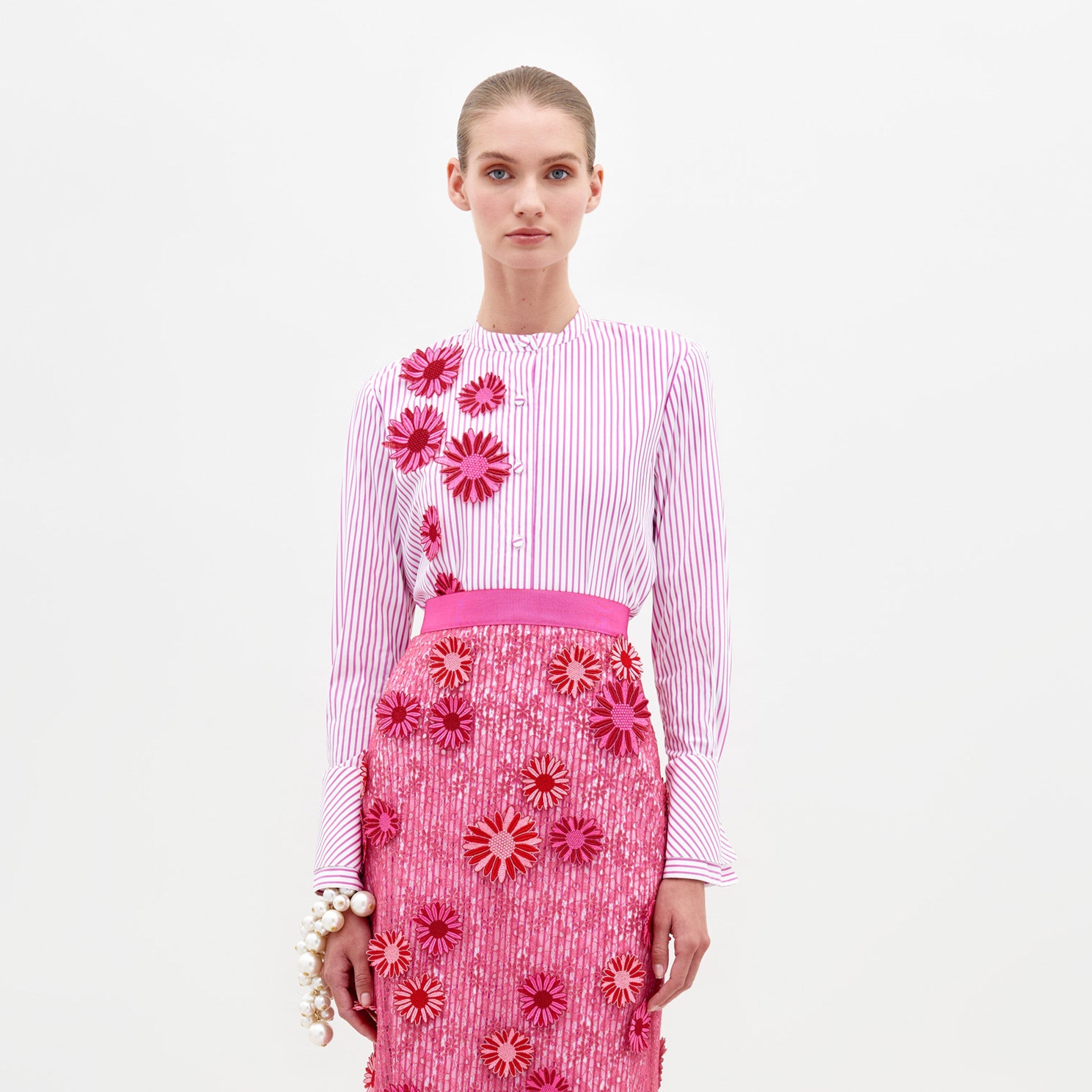 A person in the Silvina Blouse Rose Stripes adorned with floral appliqués, paired with a pink sequined skirt featuring similar floral designs, stands against a plain white background, available for pre-order now with an expected ship date in early 2025.