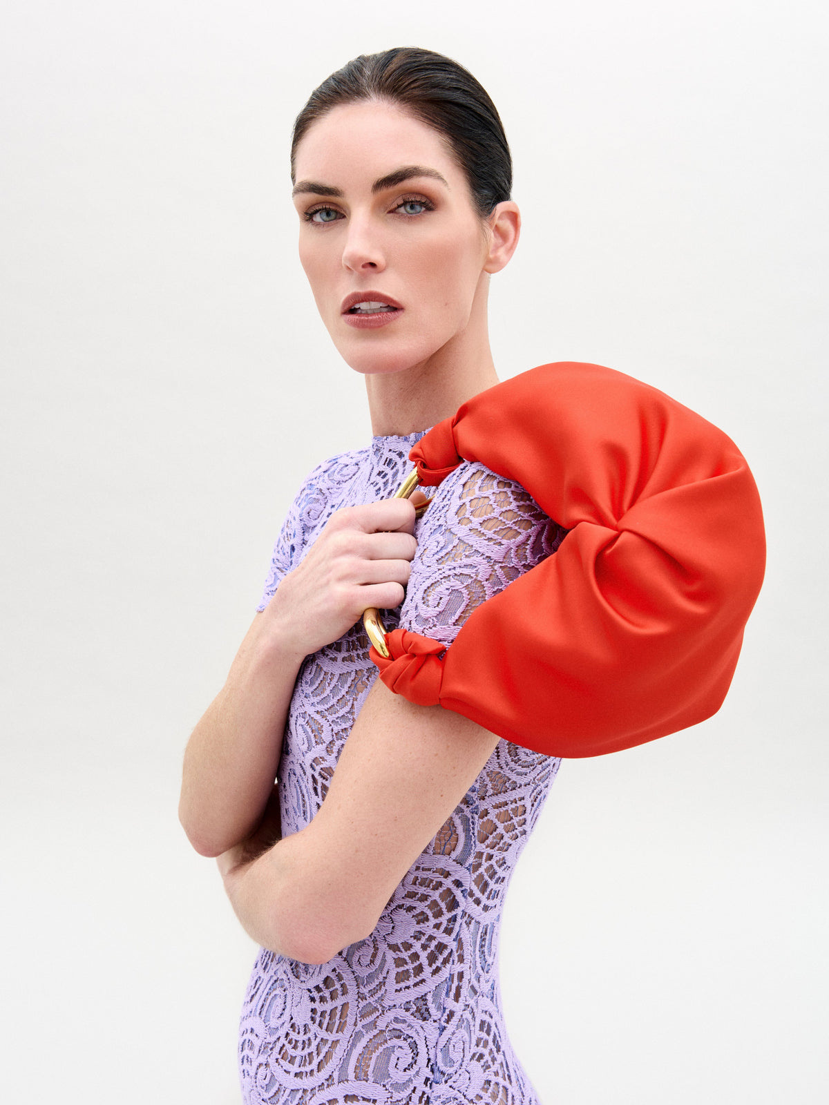 A person in a lilac lace dress holds a vibrant orange crescent-shaped Sofia Bag (Small) with a gold-tone metal handle.