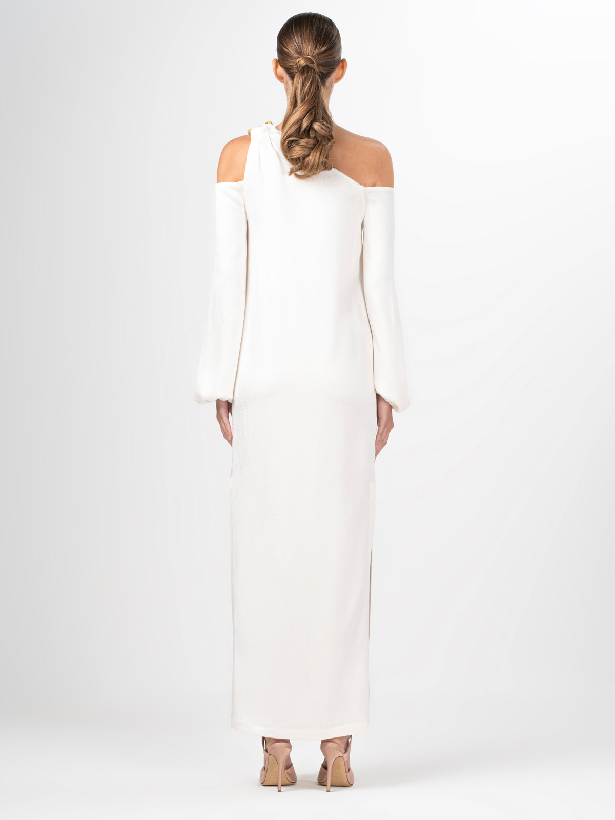 A woman with brown hair in a ponytail models the Ada Dress—an elegant, asymmetric long-sleeve design—showing off the back. She pairs it with nude high heels against a plain white background.