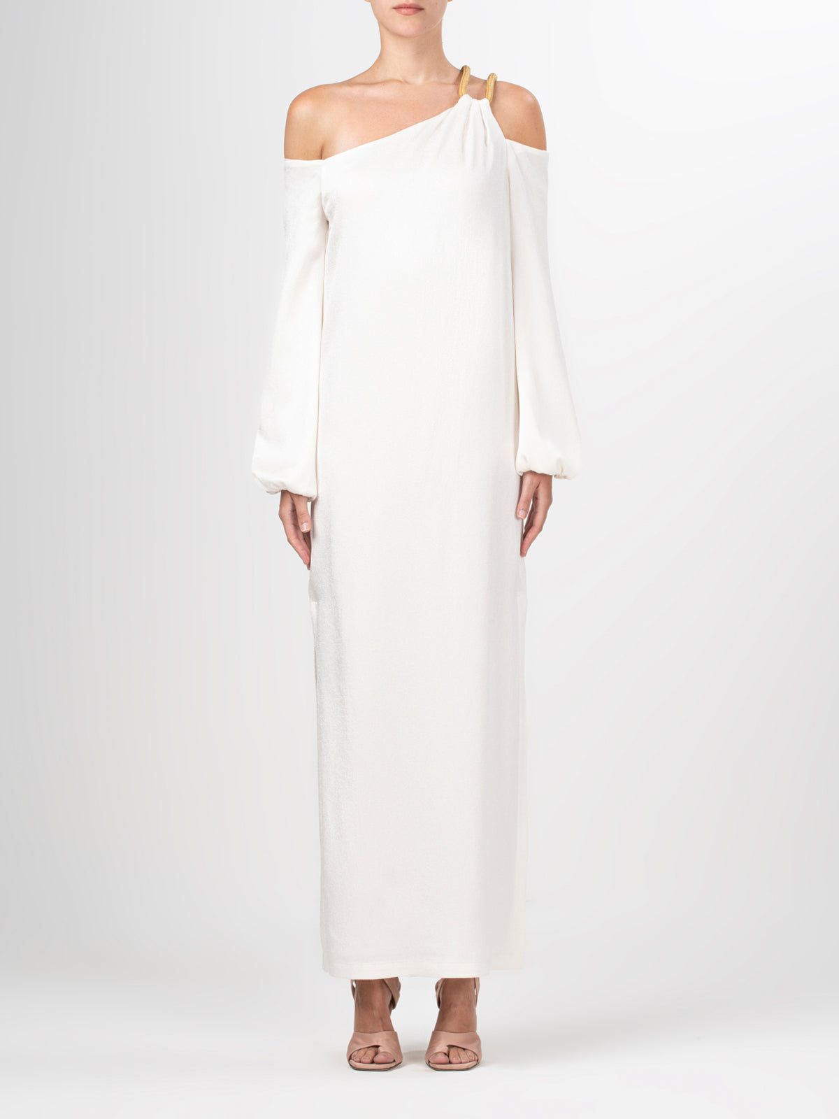 A woman models the Ada Dress—an elegant one-shoulder maxi with long sleeves and an asymmetric strap—paired with open-toe heels against a plain light background.