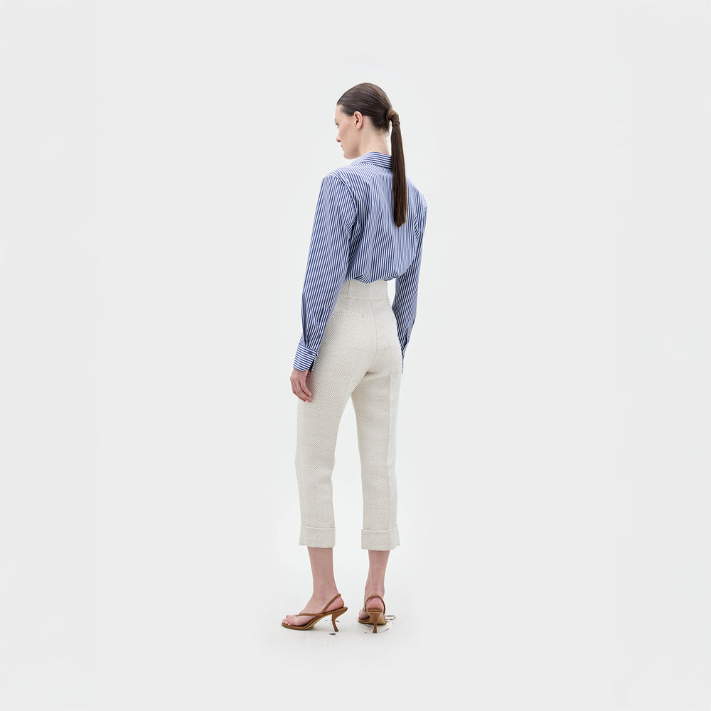 A woman stands against a plain background wearing the Karole Blouse, a blue and white striped button-down with floral embroidery made from Italian fabric, paired with high-waisted cream pants.
