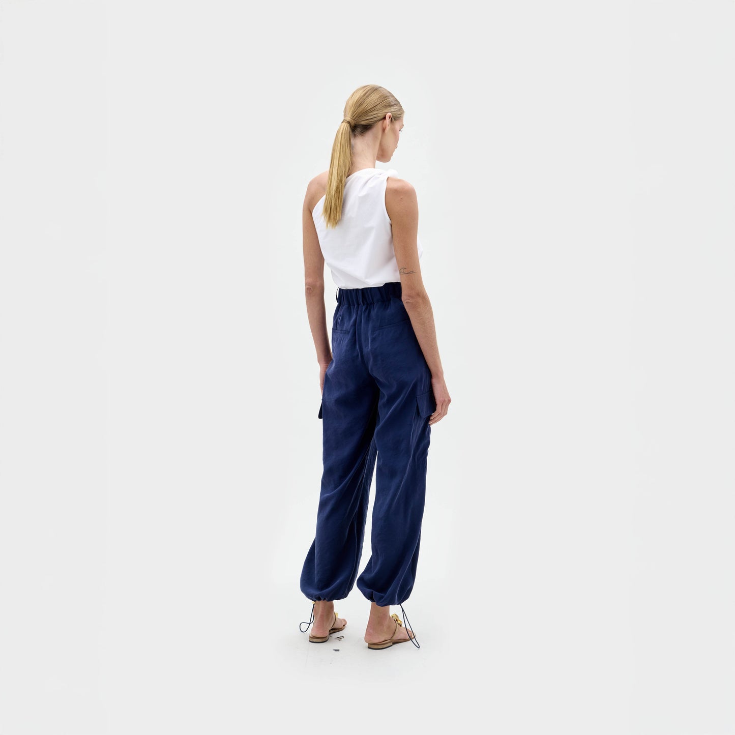 A woman stands against a plain background wearing a white one-shoulder top, brown sandals, and the Darnell Pant—navy high-waisted cargo pants made from Italian fabric.
