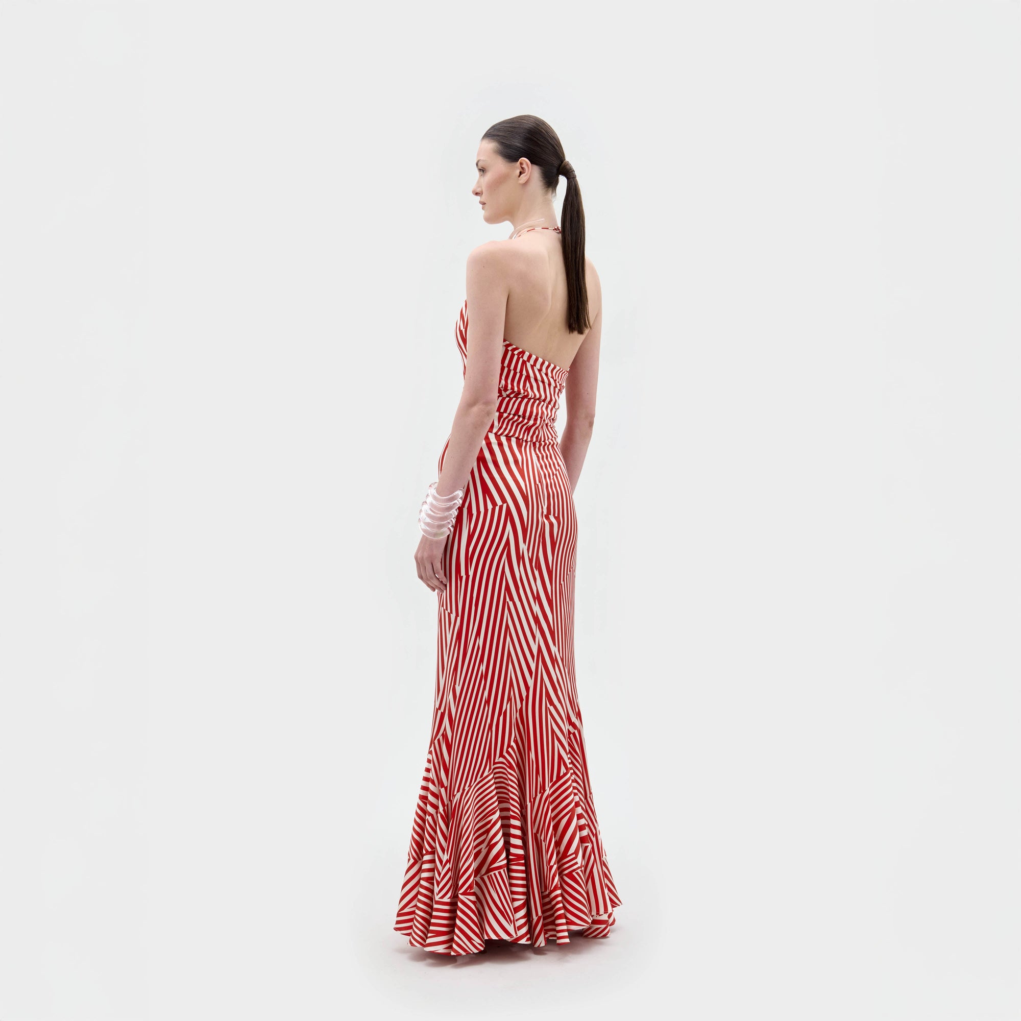 A woman stands against a plain white background wearing the Florina Skirt, a floor-length, red and white striped piece with a fitted waist and mermaid silhouette, crafted from luxurious Italian fabric.