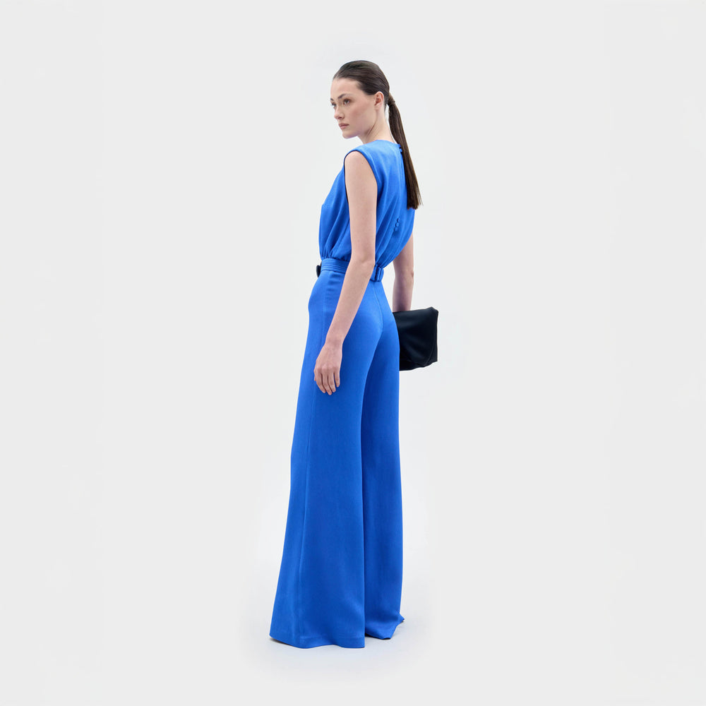 A woman stands against a plain background wearing the Noel Jumpsuit, a sleeveless bright blue wide-leg design with a gathered waist made from Italian textile, holding a black clutch.