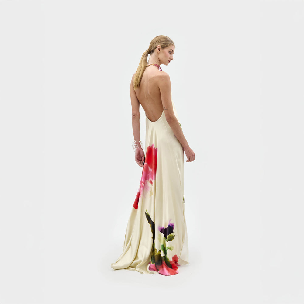 A woman stands against a plain background wearing the Jesse Dress, a sleeveless cream maxi with a colorful abstract floral print and halter neckline, paired with open-toe heels.