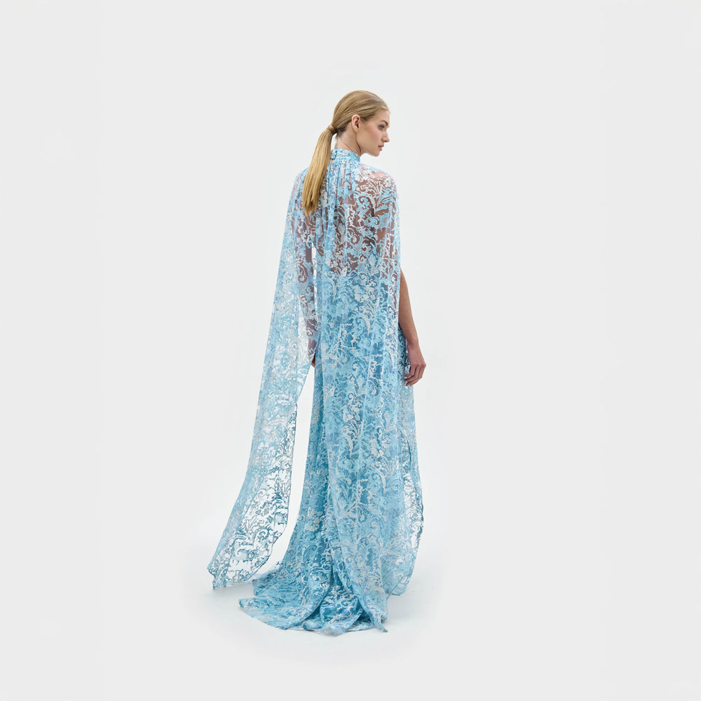 A woman models the Carol Dress—a light blue, floor-length gown made from Italian fabric, featuring a high neckline, cape shoulders, lace details, and a front slit—against a plain white background.