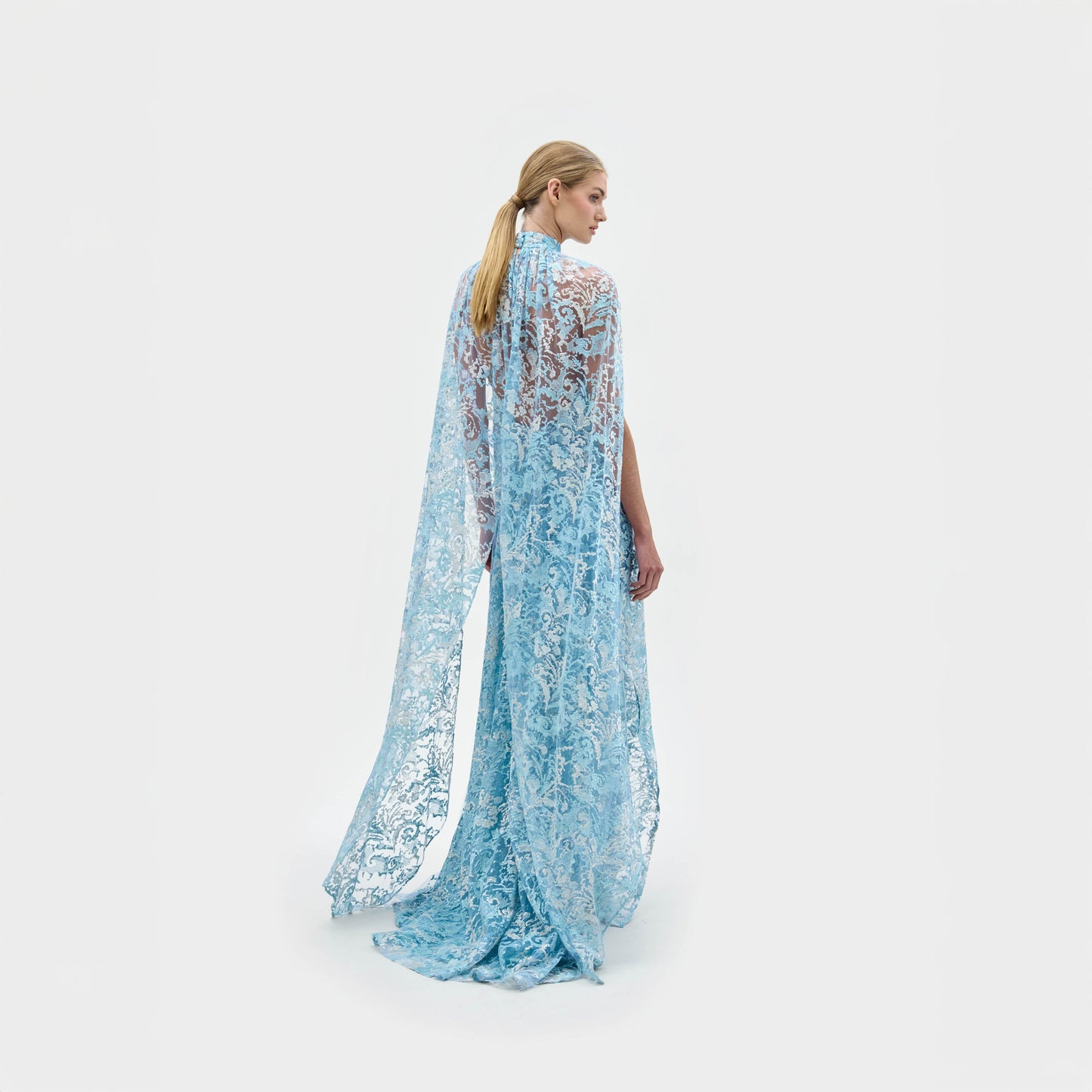 A woman models the Carol Dress—a light blue, floor-length gown made from Italian fabric, featuring a high neckline, cape shoulders, lace details, and a front slit—against a plain white background.