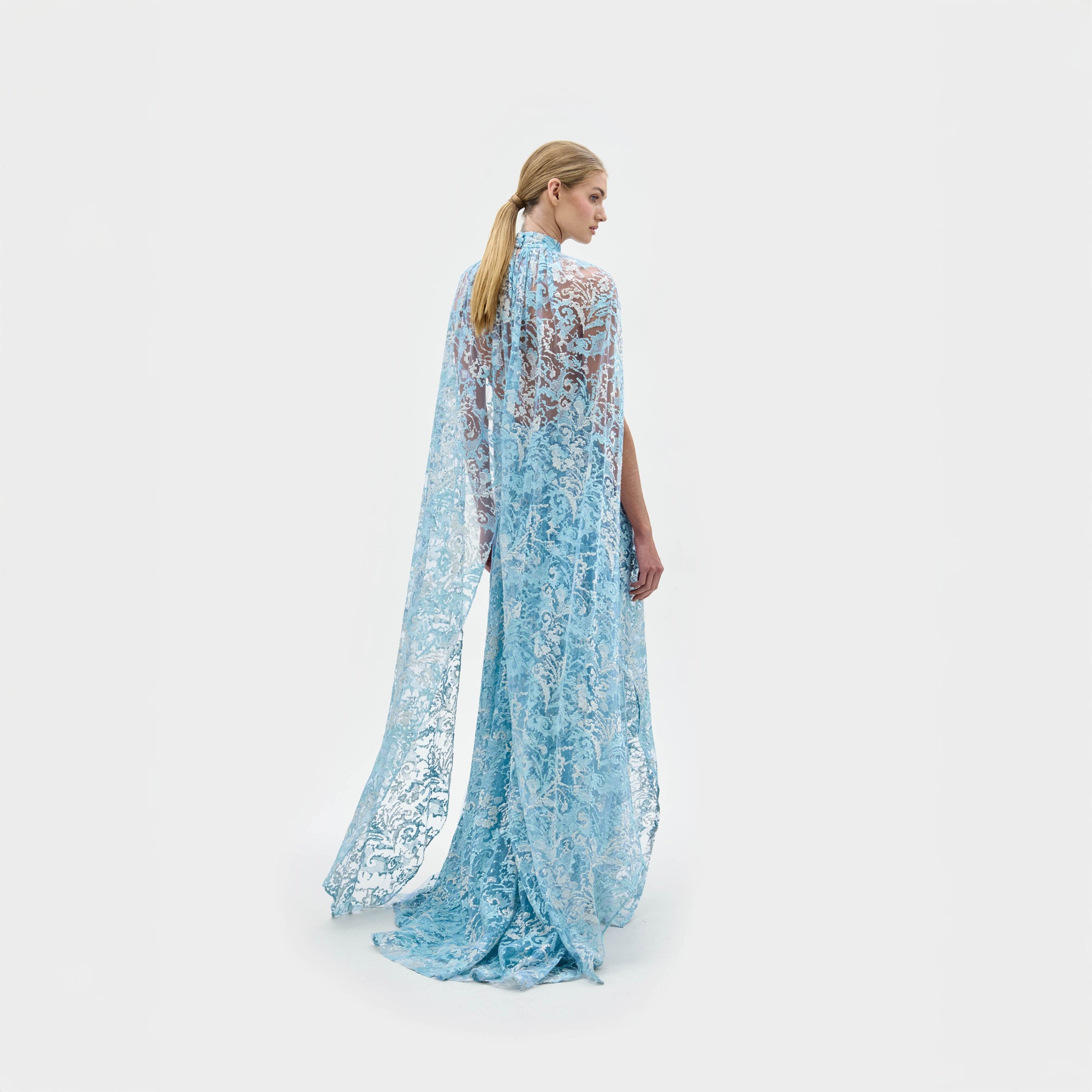 A woman models the Carol Dress—a light blue, floor-length gown made from Italian fabric, featuring a high neckline, cape shoulders, lace details, and a front slit—against a plain white background.