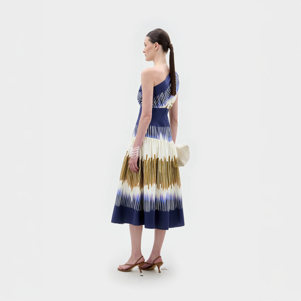 A woman stands against a plain background wearing the Catalina Dress, a one-shoulder blue and white patterned midi with an A-line silhouette, paired with a beige clutch, sandals, and stacked bracelets.
