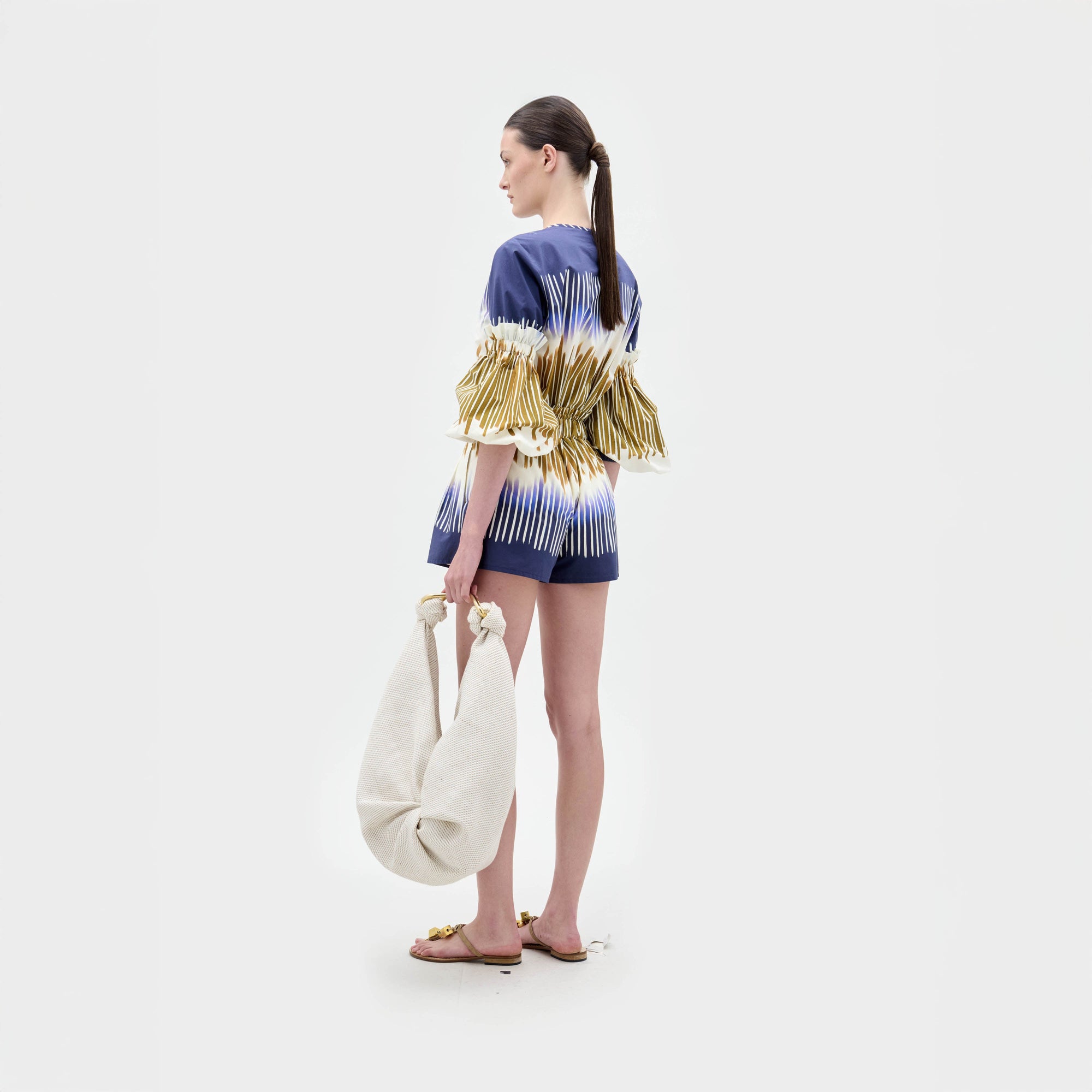 A woman stands against a plain background, wearing the Lucaya Blouse—an Italian fabric top in blue and white patterns with gold stripes, puffed sleeves, a belted waist, and elegant detailing.
