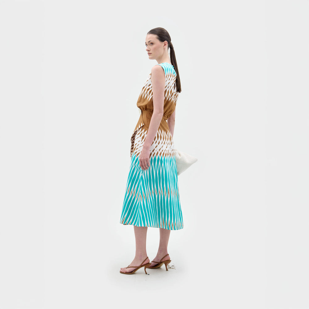 A woman stands against a white background wearing the Kendall Dress, a sleeveless turquoise, brown, and white fit-and-flare midi made from Italian fabric, paired with a large white bag and brown heeled sandals.