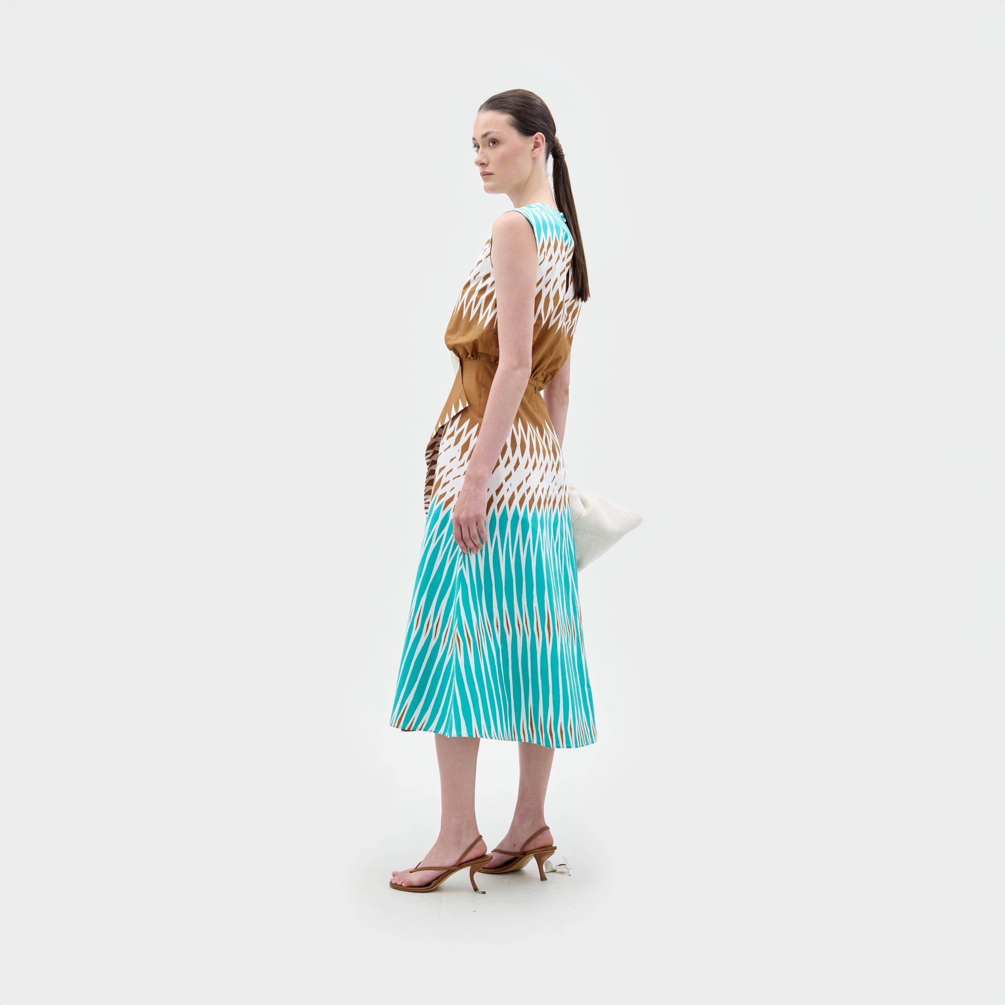 A woman stands against a white background wearing the Kendall Dress, a sleeveless turquoise, brown, and white fit-and-flare midi made from Italian fabric, paired with a large white bag and brown heeled sandals.