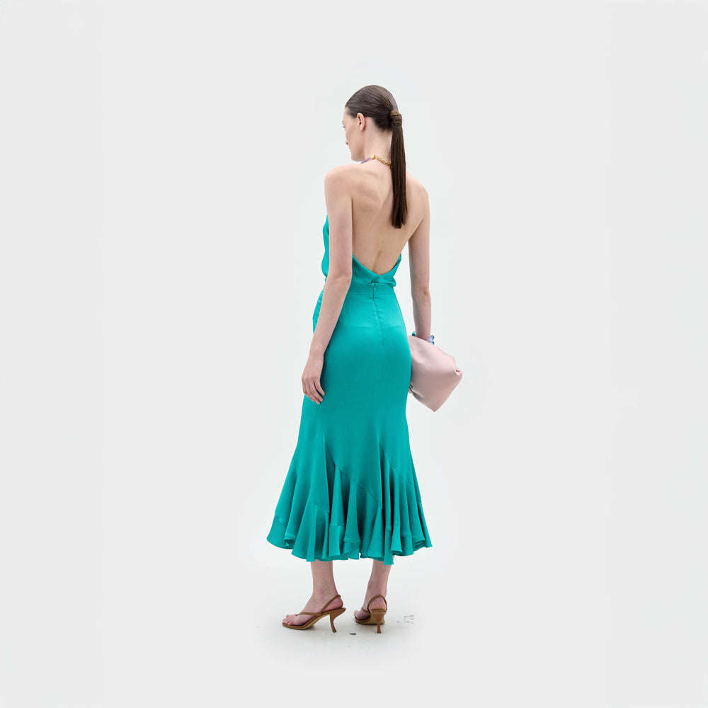A woman in the Dianela Blouse, made from luxurious Italian fabric in teal, holds a pale pink clutch against a plain white background.