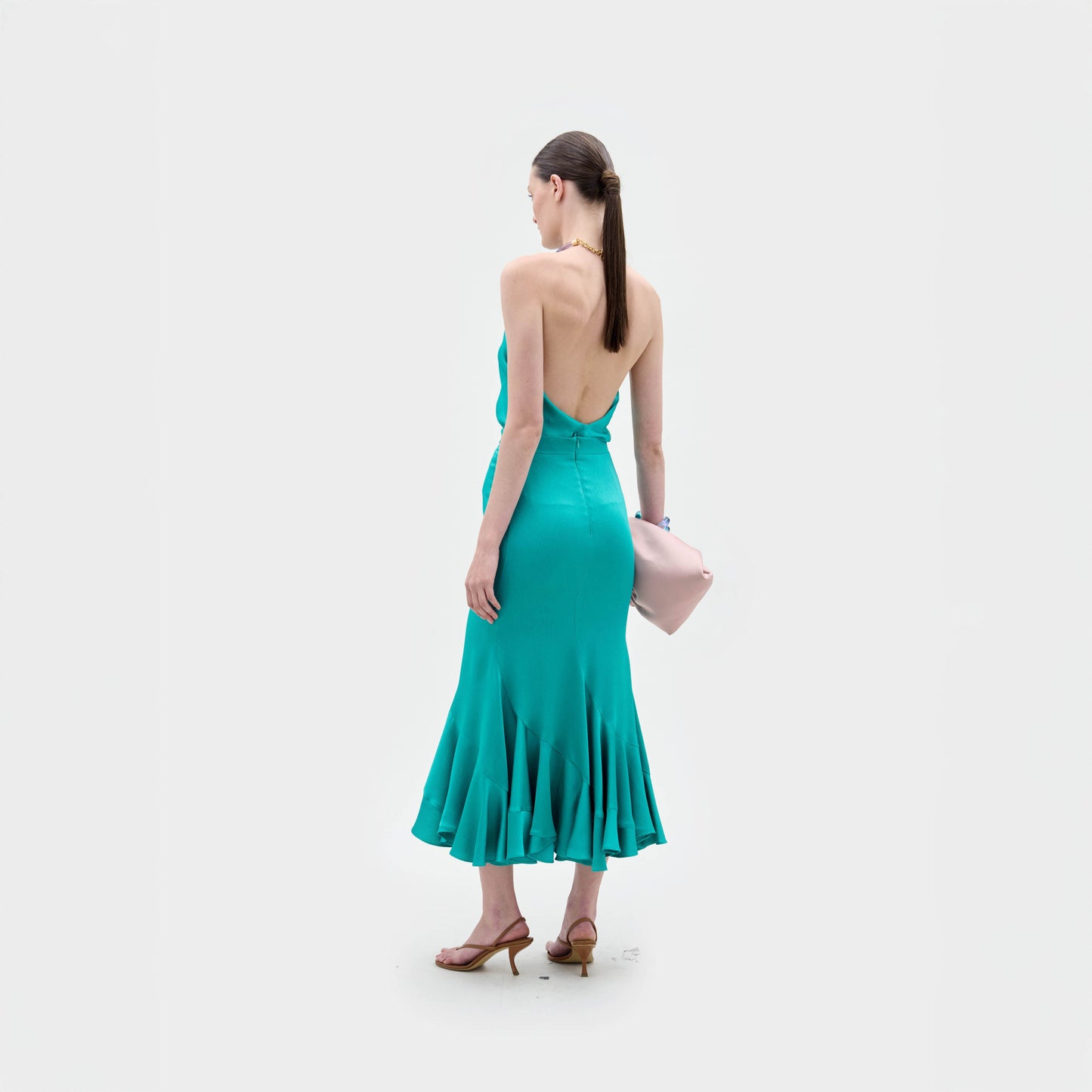 A woman stands against a plain background wearing the Marlene Skirt, styled with brown heeled sandals and holding a large pale pink clutch.