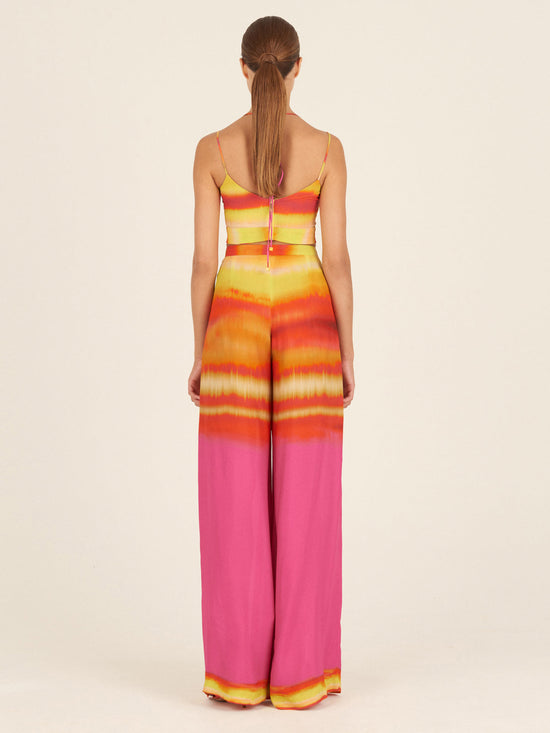 A woman is seen from behind wearing a colorful jumpsuit with wide-leg trousers in a Benedetto Pant Fuschia Lime Stripes pattern of yellow, orange, and pink, standing against a neutral background.