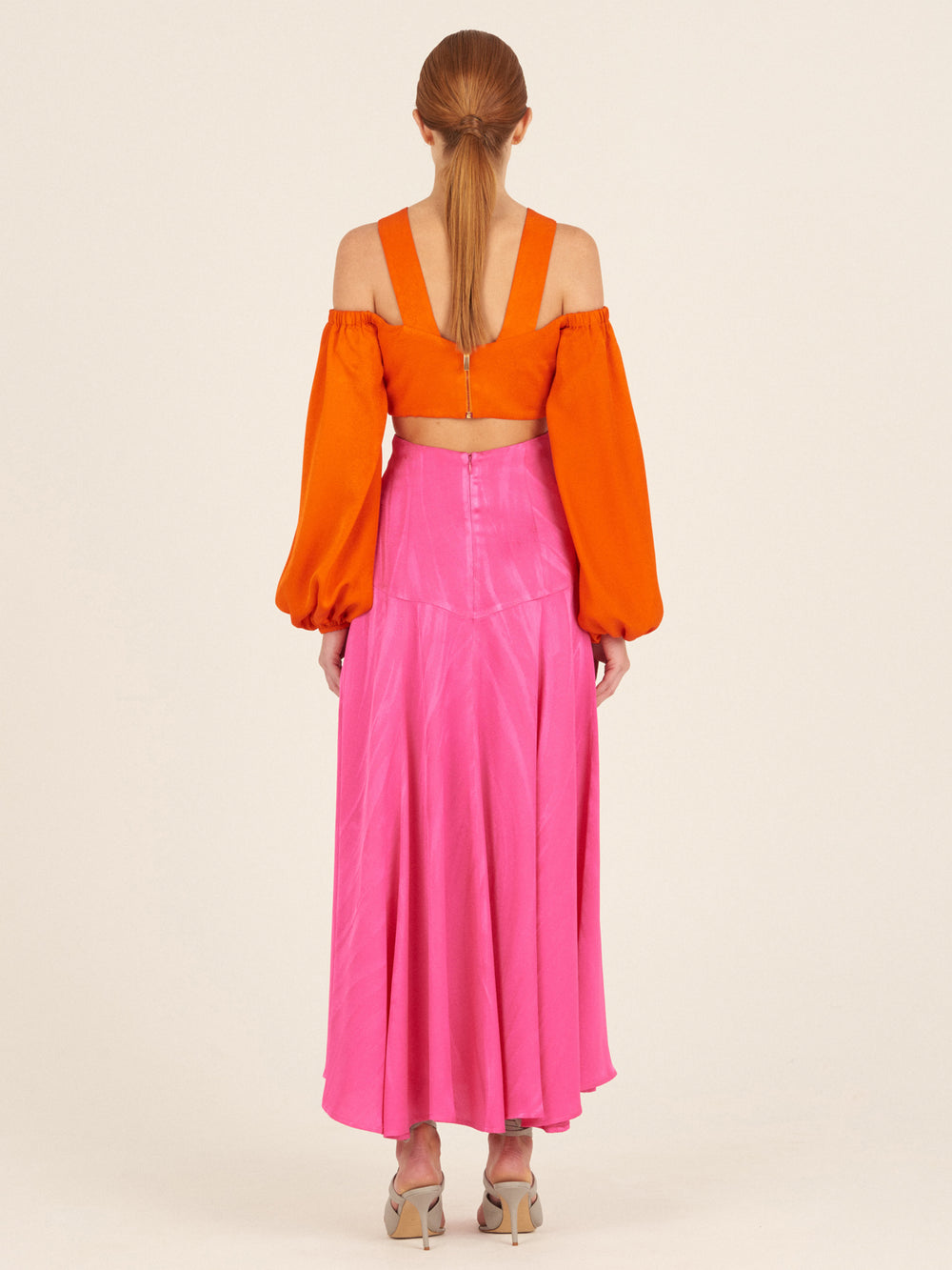 A woman faces away wearing the Carole Top in bright orange, a cropped off-the-shoulder style with billowing sleeves, paired with a high-waisted, ankle-length pink jacquard panel skirt and beige heeled sandals.