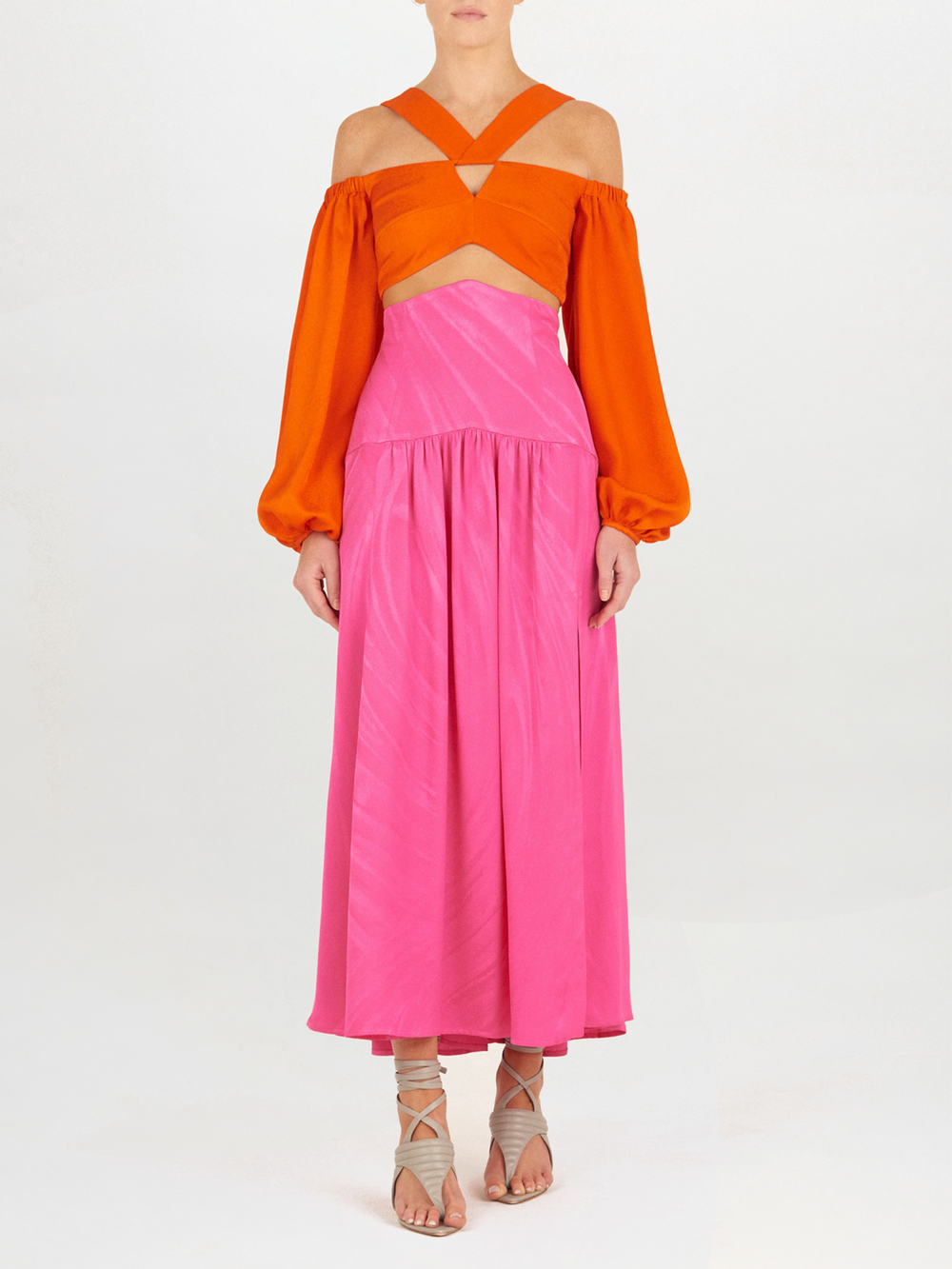 A person wears the Carole Top Bright Orange—an off-the-shoulder, crisscross cropped top with billowing sleeves—paired with a high-waisted pink skirt and beige strappy sandals against a plain white background.