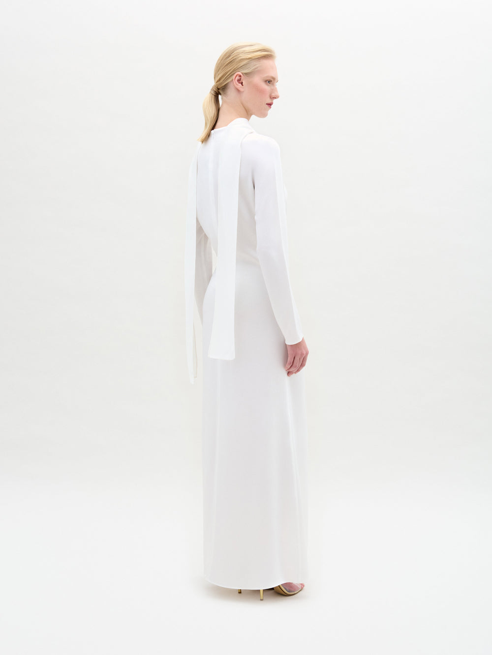 A woman stands against a white background wearing the Sue Dress, a long-sleeved satin maxi with a twist detail at the waist, paired with gold bracelets and gold open-toe heels.