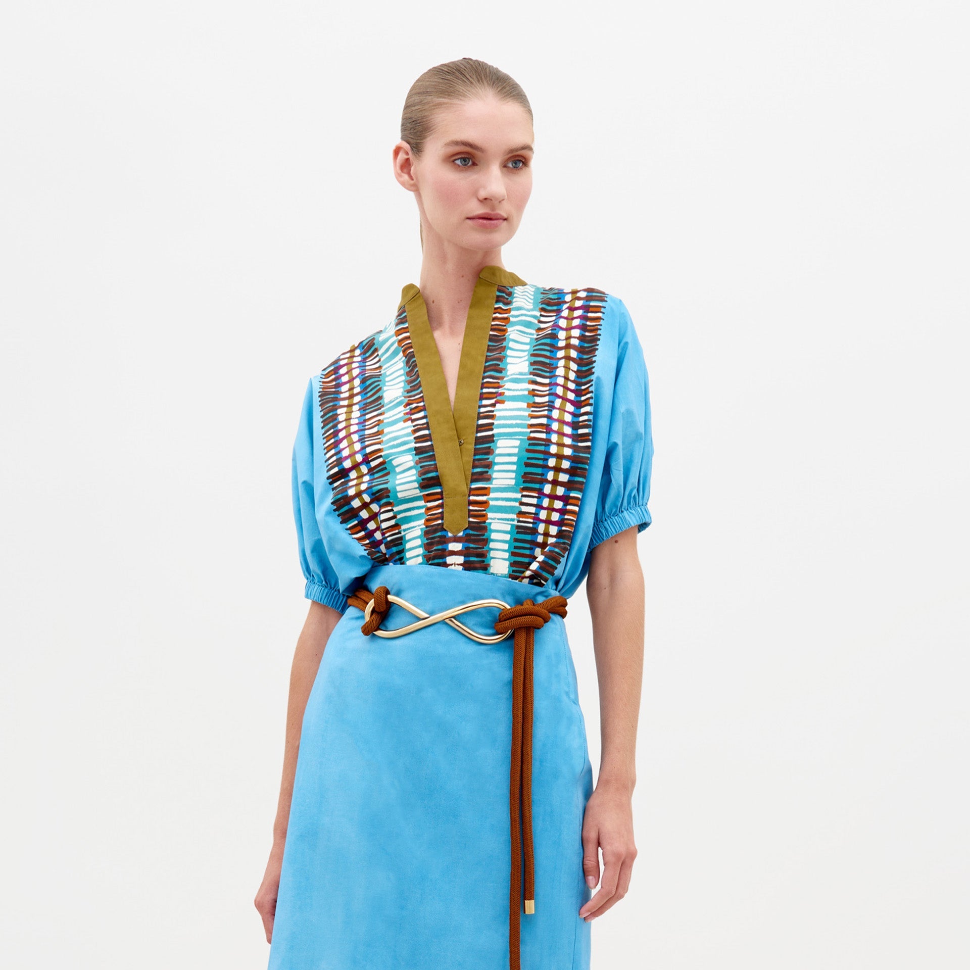 A woman wears the Susanne Blouse in Aqua Olive Plaid, featuring a multi-colored patterned V-neck and a knotted brown belt, as she stands against a plain white background. This elegant outfit is available for pre-order now and will ship by February 15th, 2025.