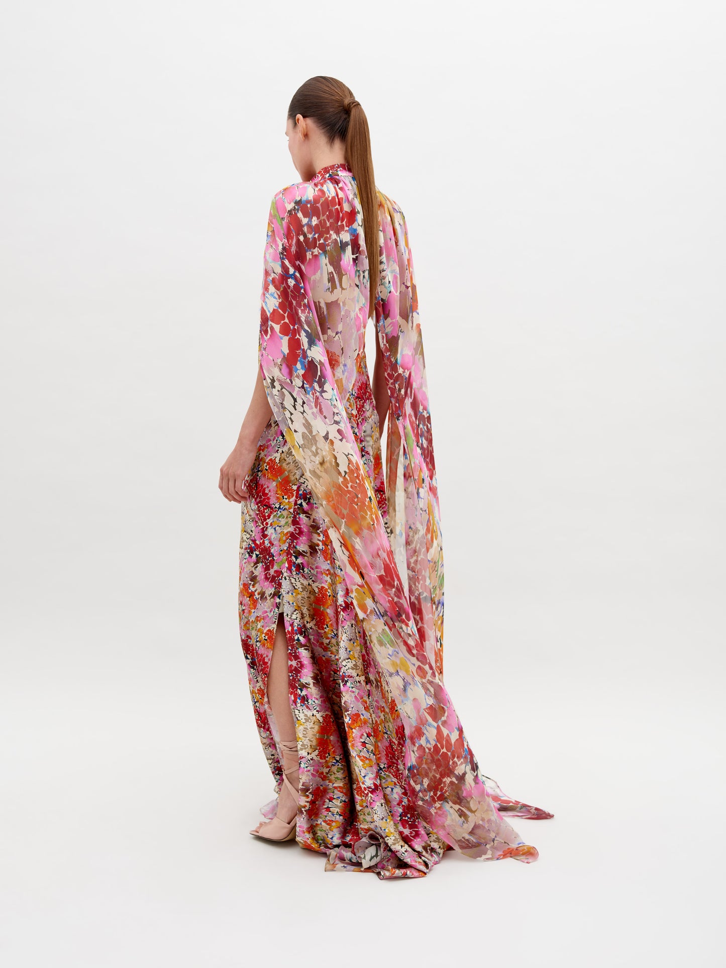 A person stands against a white background wearing a colorful, floral-patterned long gown with a cape, and strappy sandals. The Sabina Dress Multicolor Animal Print is available for pre-order now; order by November 15th, 2024 to ensure timely delivery.