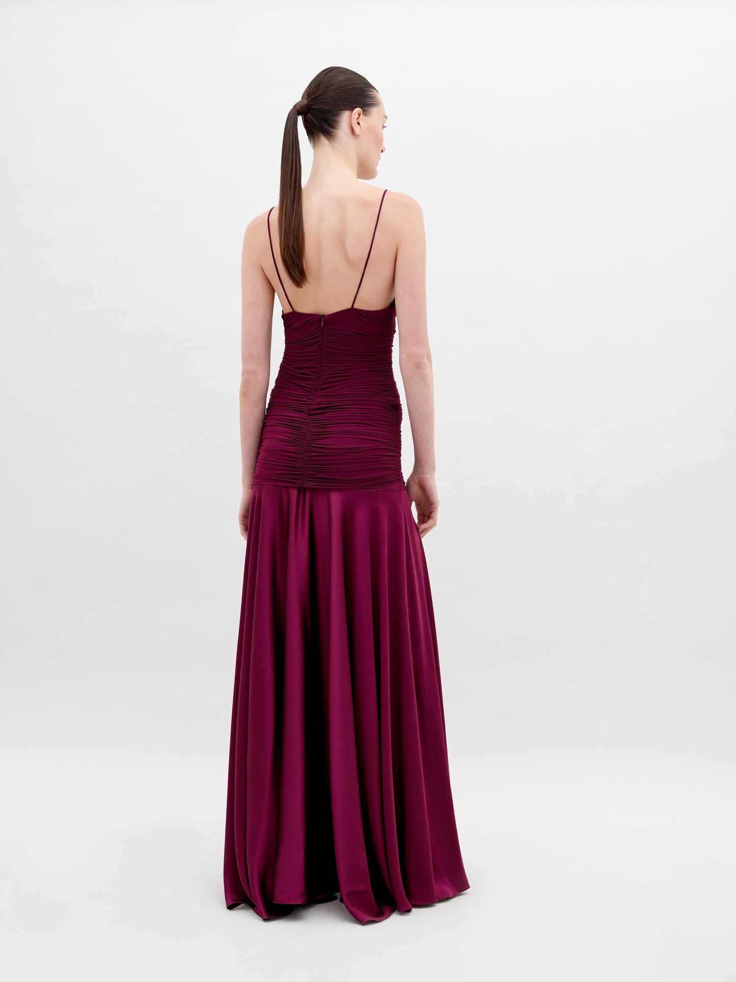 A woman stands in a long, sleeveless, deep red Sana Dress Burgundy with ruching and floral detailing on the bodice. She has her hair pulled back and is looking slightly to the side. The background is plain white. Pre-order now for a ship by date of November 15th, 2024.