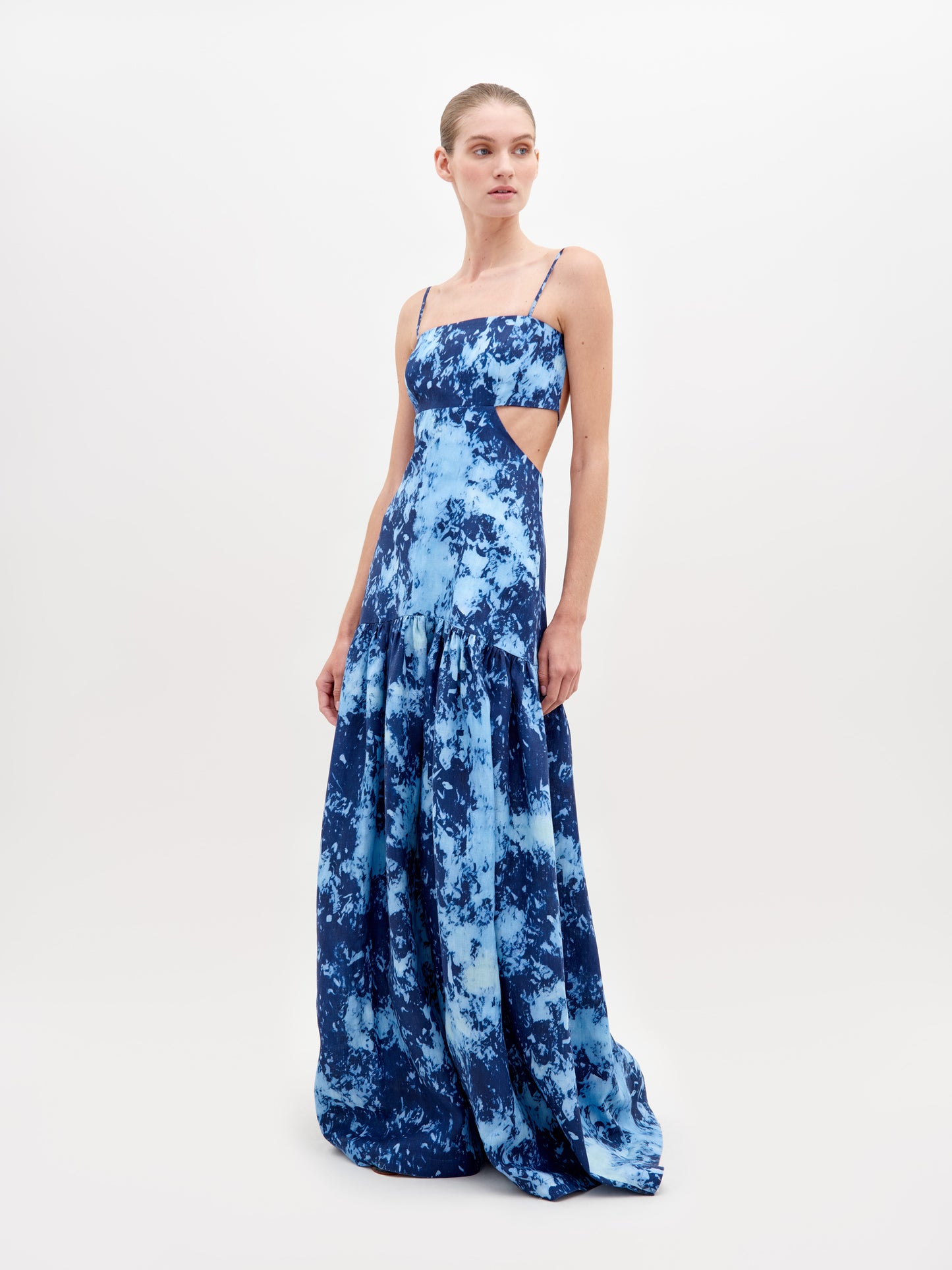 A woman stands wearing a long, blue, tie-dye sleeveless dress with thin straps and side cutouts, posing against a plain white background. The Shannon Dress Indigo is available for pre-order now and will ship by November 15th, 2024.