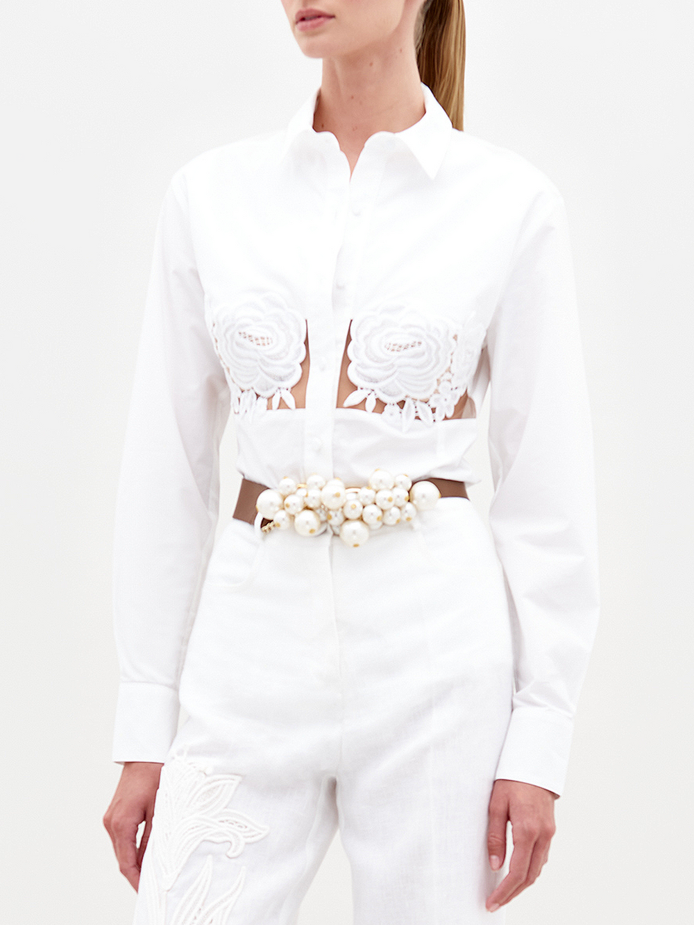 A woman wears the Sigrid Blouse—white cotton with floral appliqué on the chest—and high-waisted white pants, styled with a pearl belt to showcase refined Italian craftsmanship. Her hair is pulled back for an elegant look.
