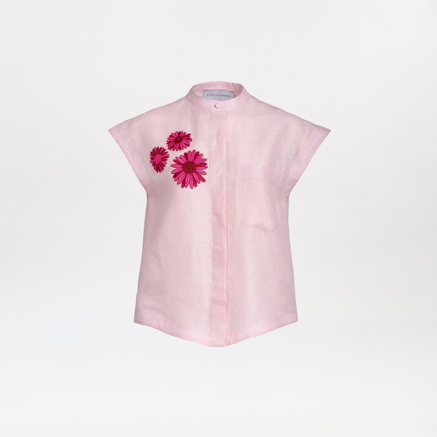 The Adele Blouse is a light pink, short-sleeve top crafted from 100% Italian cotton, featuring an elegant mandarin collar and three vibrant pink embroidered flowers on the left chest. The blouse is set against a plain white background.