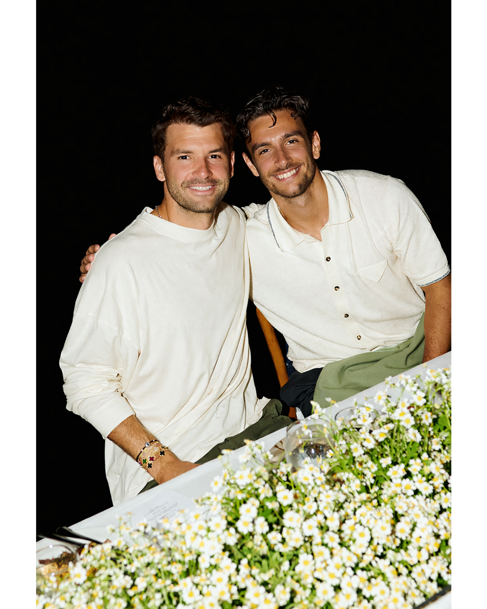 Two men in white shirts standing next to a table with flowers on a dark background