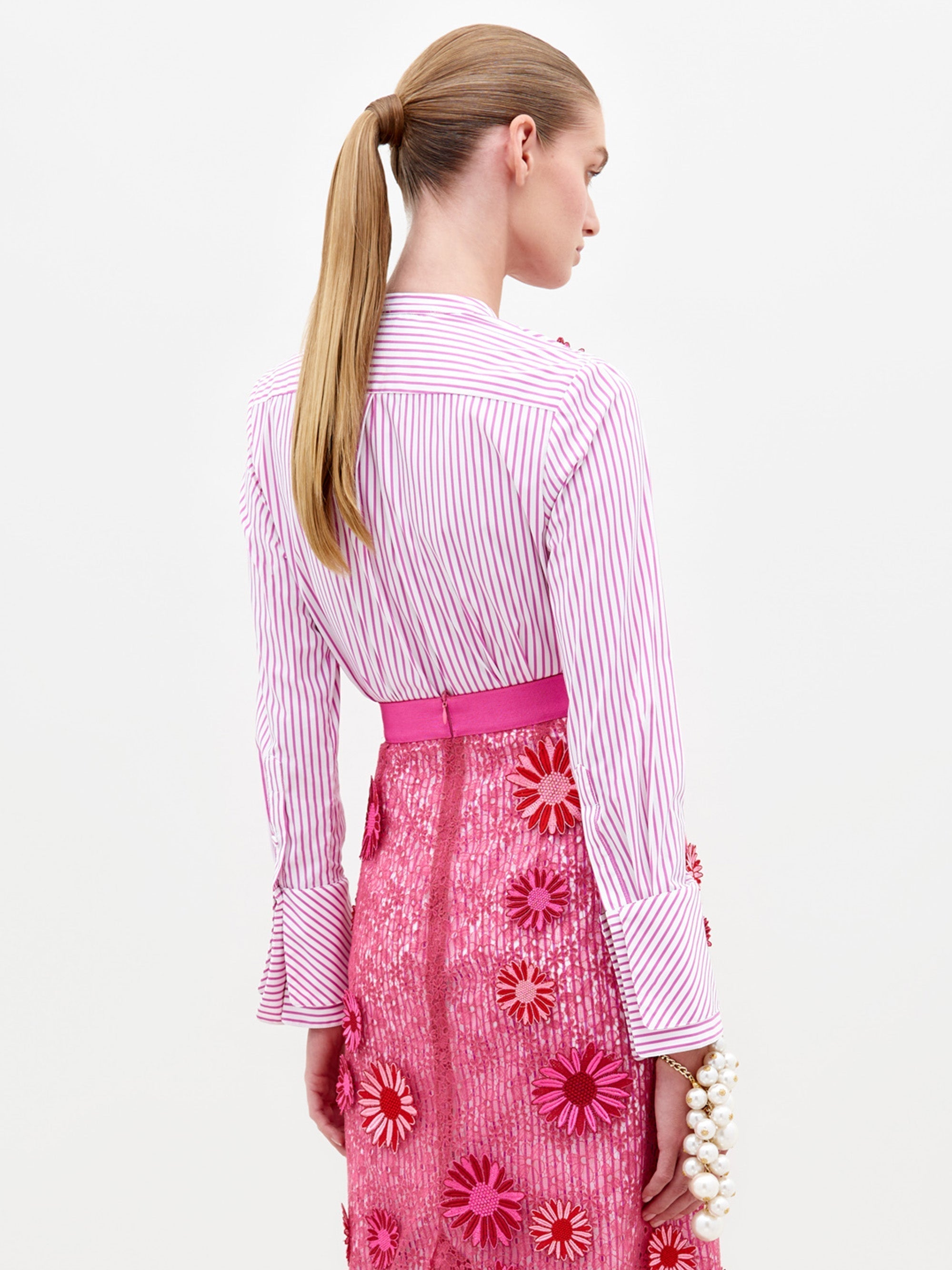 A person in the Silvina Blouse Rose Stripes adorned with floral appliqués, paired with a pink sequined skirt featuring similar floral designs, stands against a plain white background, available for pre-order now with an expected ship date in early 2025.