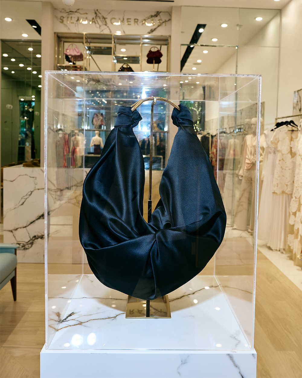 A large, curved black fabric sculpture is displayed in a transparent glass case within a clothing store. The case is mounted on a marble pedestal.