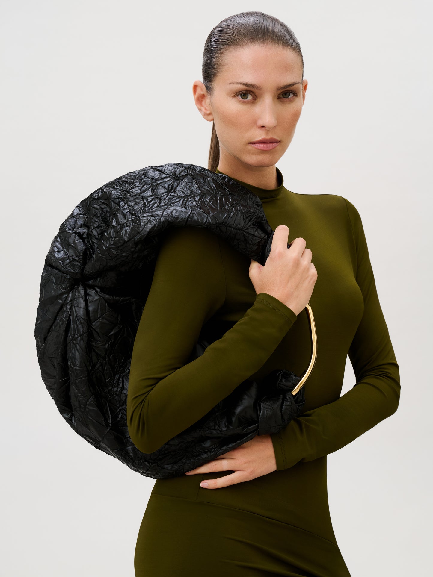 A person in an olive green, long-sleeved dress stands holding the Sofia Bag (Maxi) with a gold-toned handle over one shoulder, looking directly at the camera against a plain light background.