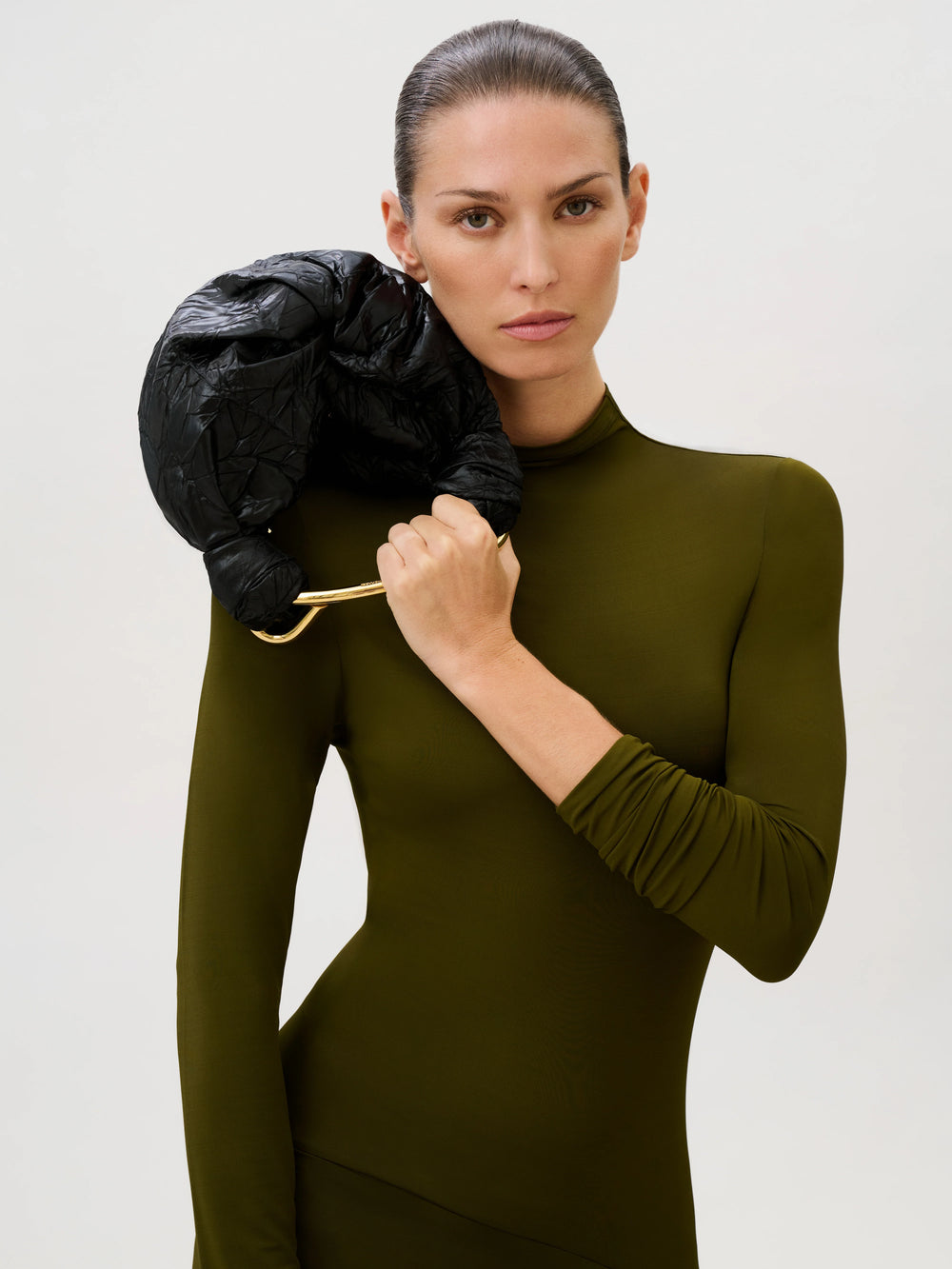 A woman in a long-sleeved olive dress holds a small, black, gathered Sofia Bag (Small) with an Italian fabric design and a gold-toned metal handle.