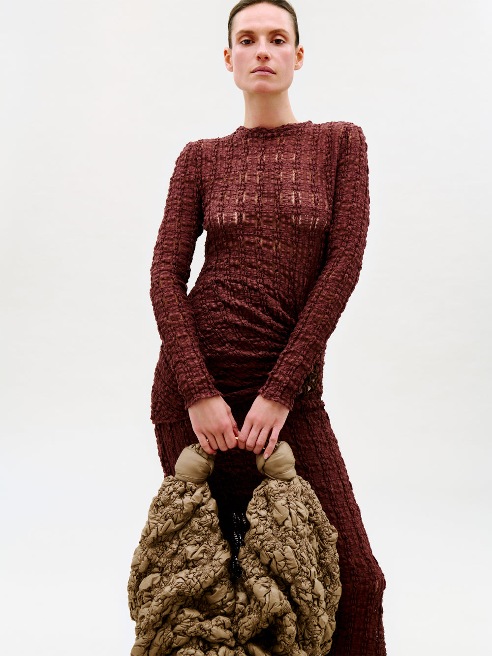 A person in a textured burgundy dress stands against a plain background, holding the Sofia Bag (Maxi), its gold-toned handle gleaming as they grip the Italian-crafted piece with both hands in front of them.