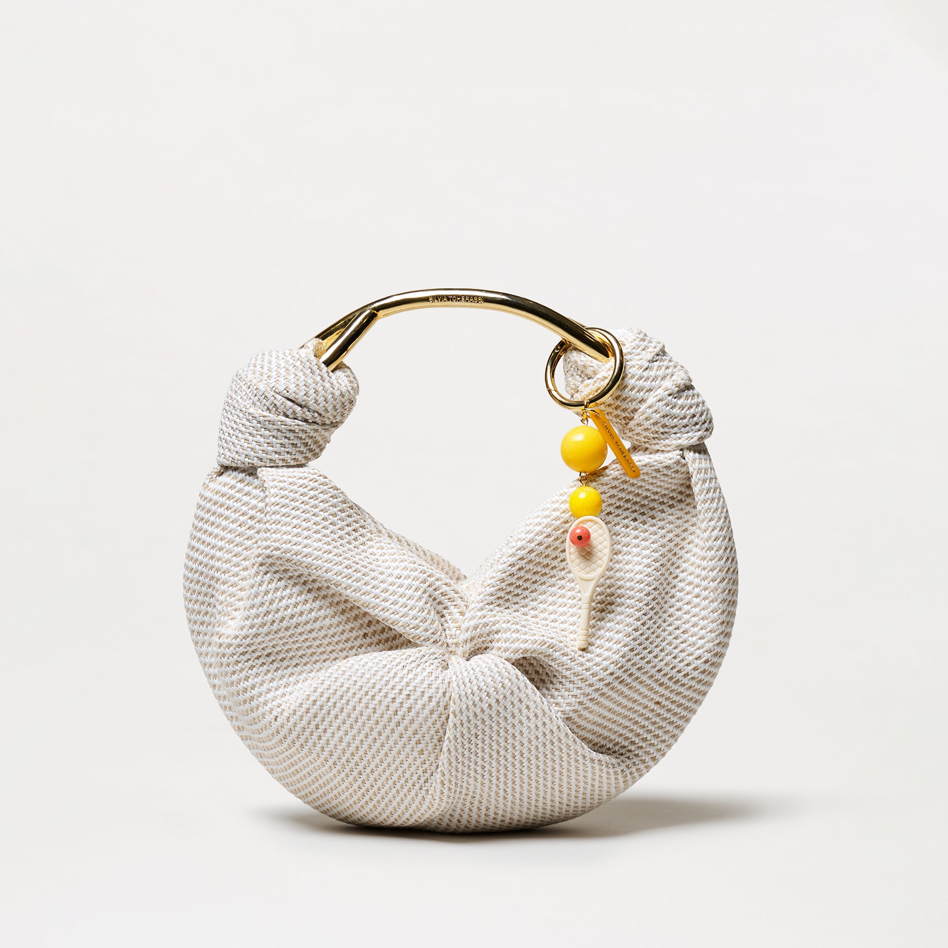 The Deuce Charm is a crescent-shaped handbag featuring textured white and gold fabric, a curved metallic gold handle, and a yellow beaded keychain with a small decorative spoon attached.