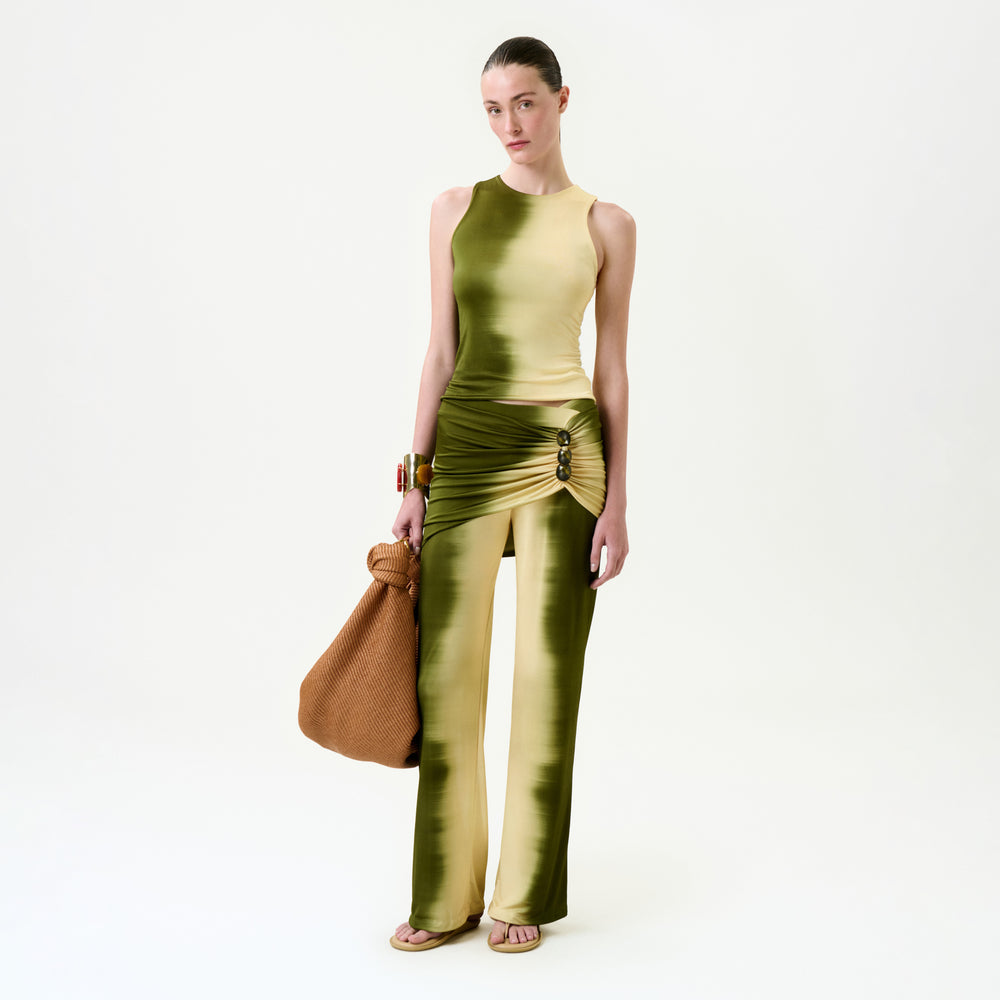A person wearing the Sonu Pant, a slim fit green and yellow gradient pant with a side slit, holds a large brown textured bag while standing against a plain white background.