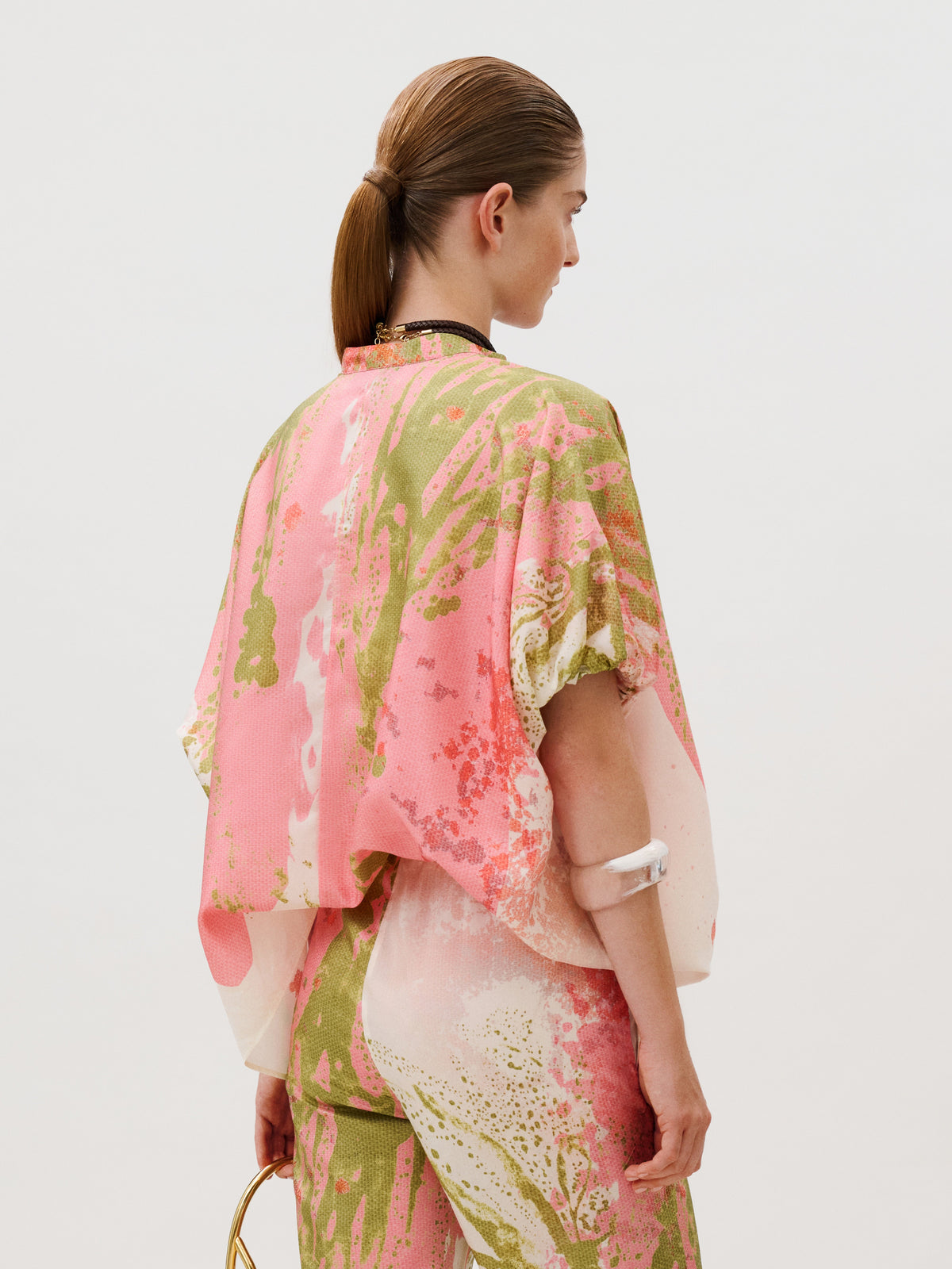 A person seen from the back wearing a Susane Blouse, a loose, short-sleeved silk top with an abstract pink and green pattern.
