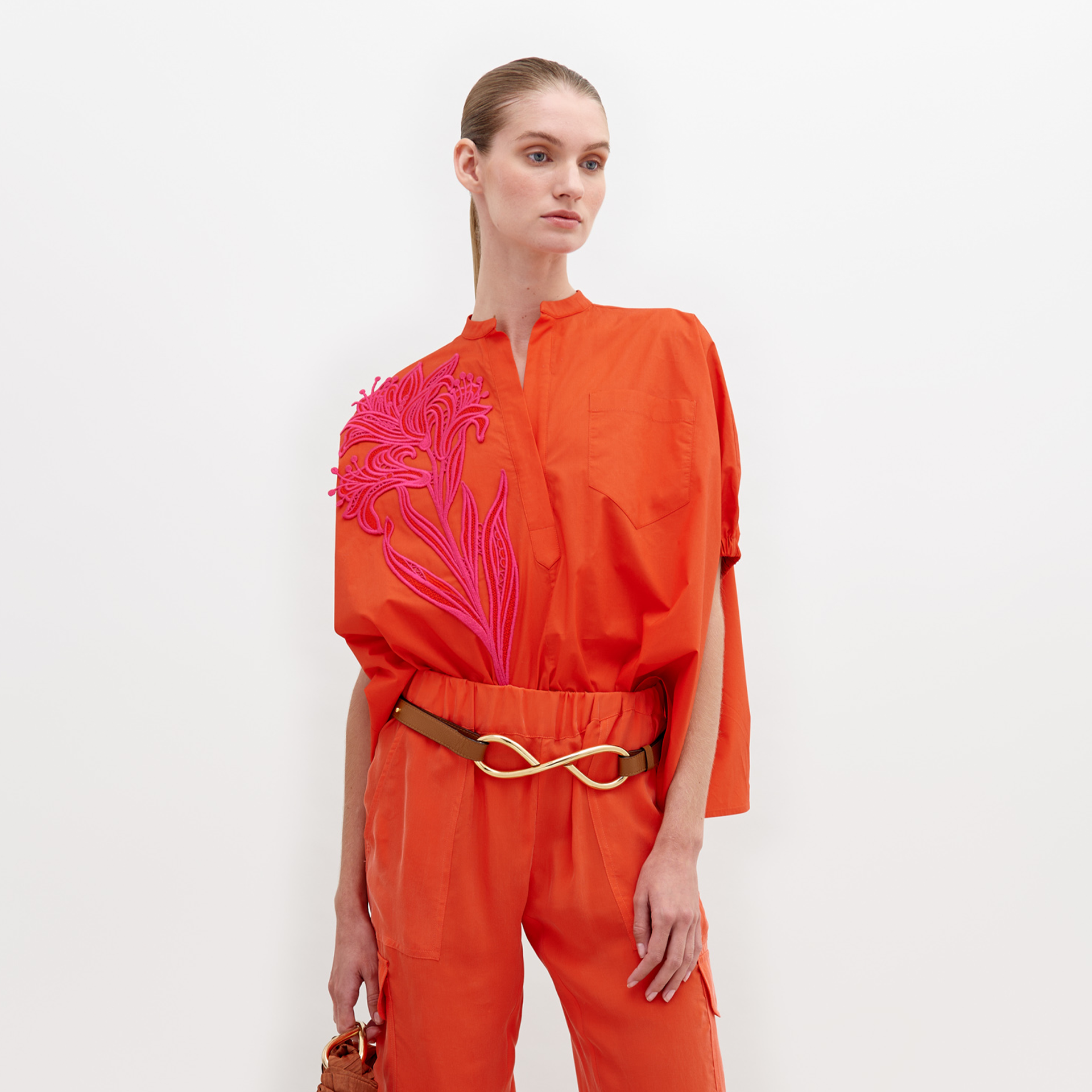 A woman stands against a white background, wearing an orange outfit with a pink floral pattern on the upper left side and a gold belt. The Susanne Blouse Scarlet is available for pre-order until November 15th, 2024.