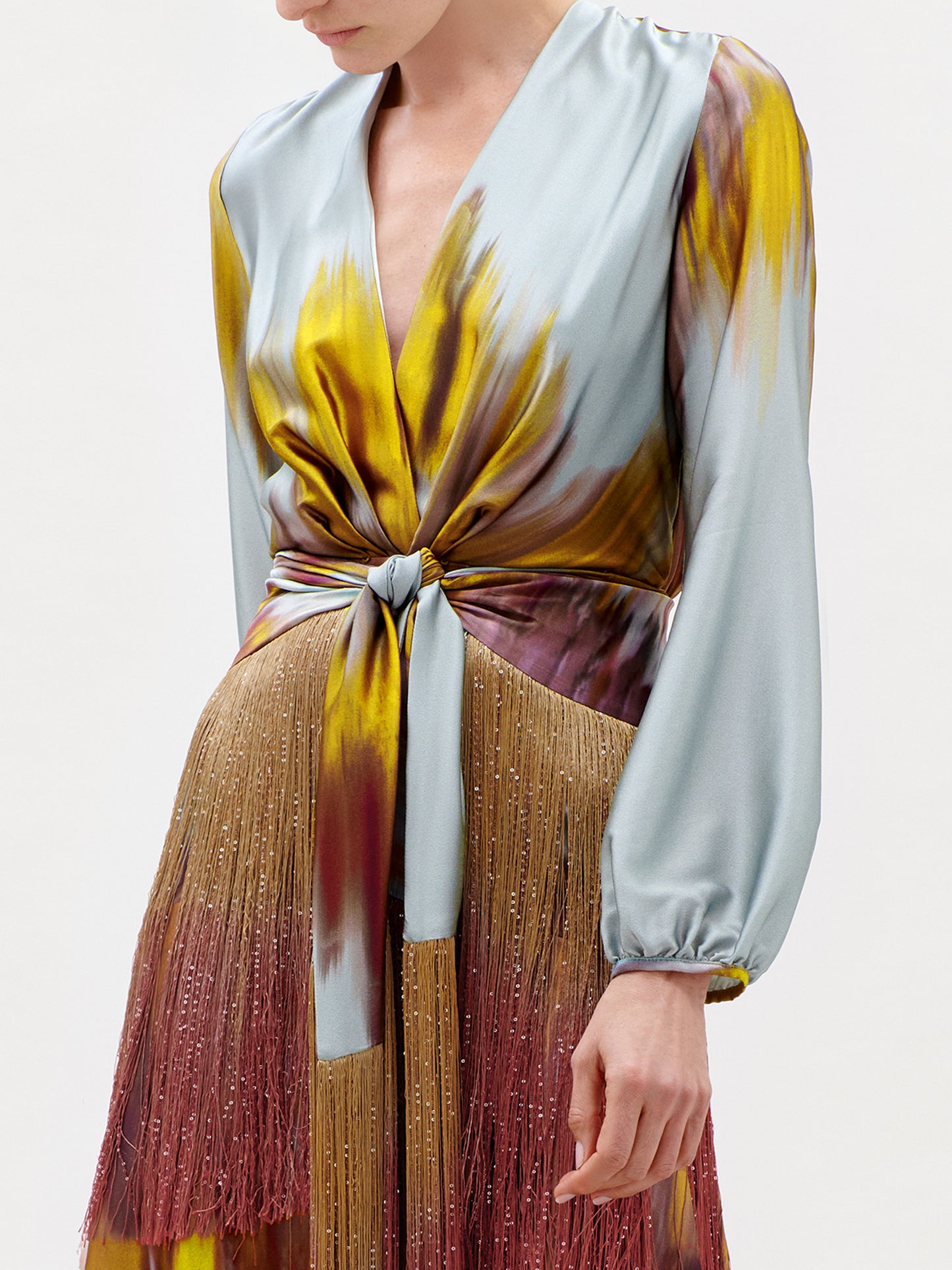 A woman wearing the Sutton Dress—a luxurious Italian silk maxi featuring long sleeves, a deep V-neckline, a knot at the waist, and a striking multicolored pattern in blue, yellow, and burgundy. This stunning garment is finished with fringe details along the skirt.