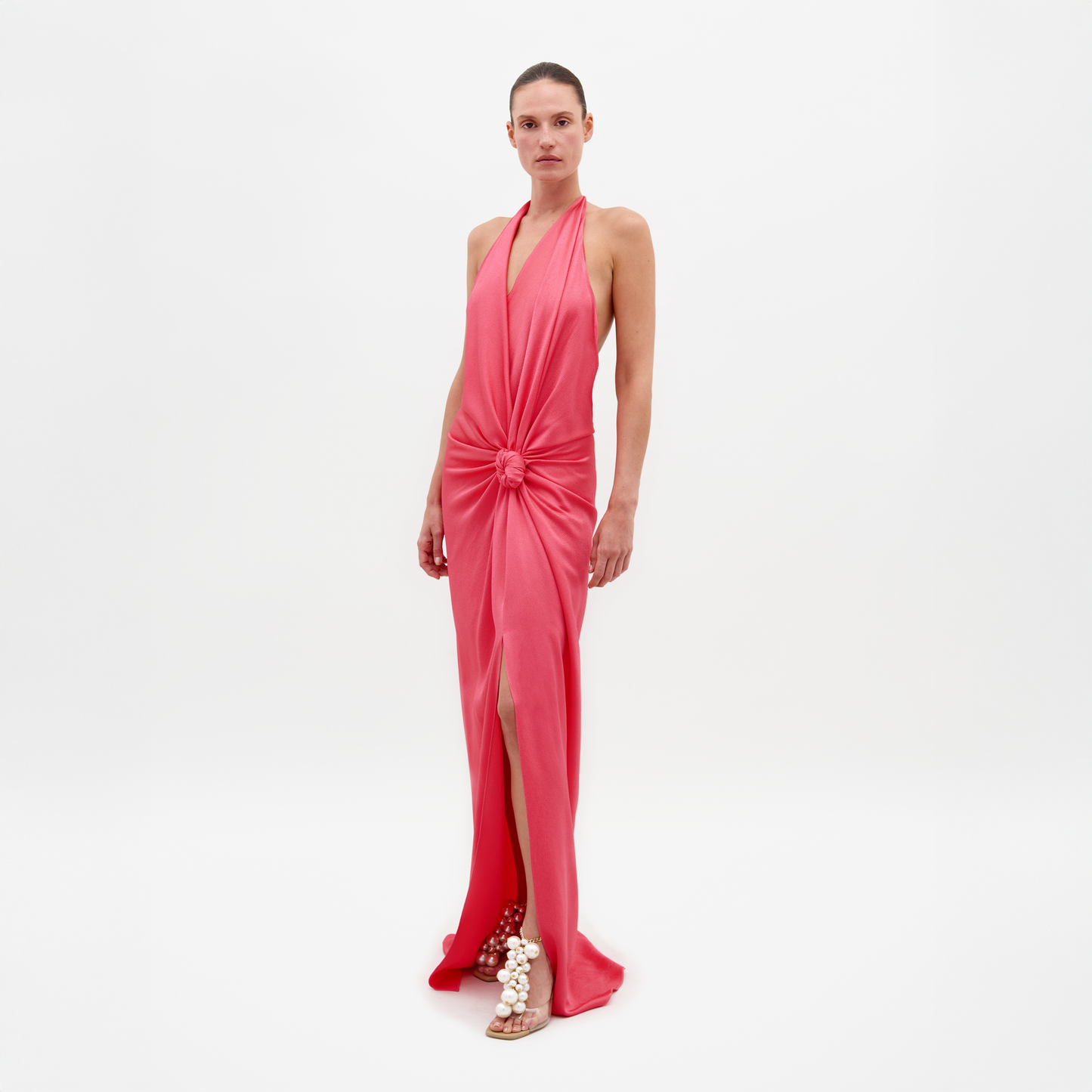 A person wearing the Torgiano Dress Peony, which features a sleeveless, V-neck design with a knot detail at the front and a slit, standing against a plain white background. Pre-order now to ensure delivery by February 15th, 2025.