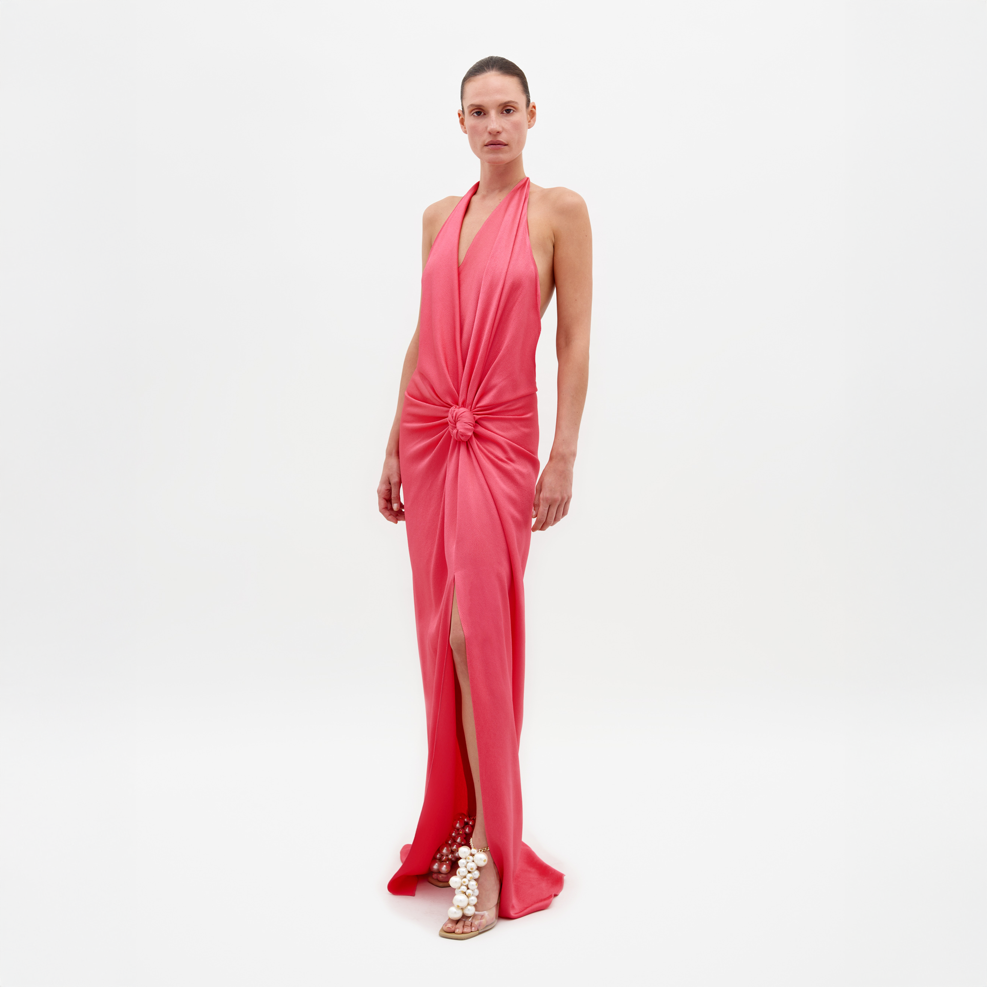 A person wearing the Torgiano Dress Peony, which features a sleeveless, V-neck design with a knot detail at the front and a slit, standing against a plain white background. Pre-order now to ensure delivery by February 15th, 2025.