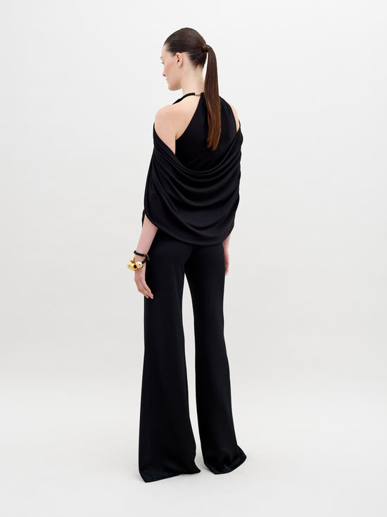 A person stands against a plain background wearing a stylish, sleeveless black top with draped fabric and Ovidia Pant Black, accessorized with gold-toned bracelets and sandals. Available for pre-order now, this ensemble will ship by date in early 2024.