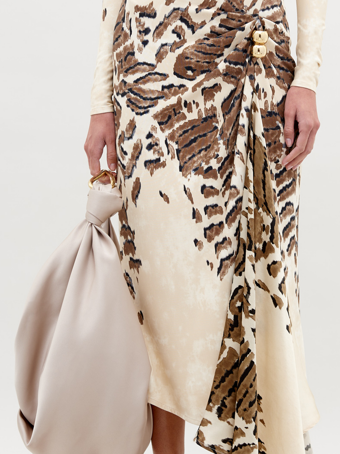 A woman in an off-shoulder, floor-length Tahia Dress Beige Abstract Animal Print stands against a plain background, holding a large beige bag in one hand. She is wearing gold sandals and a gold choker. Mark your calendars for November 15th—this stunning look is available for pre-order ahead of its 2024 launch.