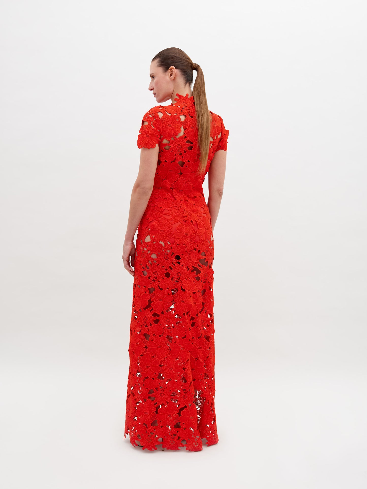 Person wearing a floor-length, red lace Taiana Dress Rouge with a high neckline and short sleeves, standing against a plain white background. Pre-order now to ensure delivery by the November 15th, 2024 ship-by date.