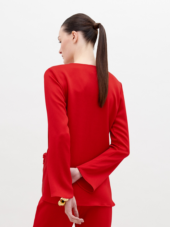 A woman stands in a red outfit with a knot detail on the top. She has her hair pulled back and stares directly into the camera against a plain white background. Available for pre-order now, the Tayra Blouse Rouge will have a ship date of November 15th, 2024.