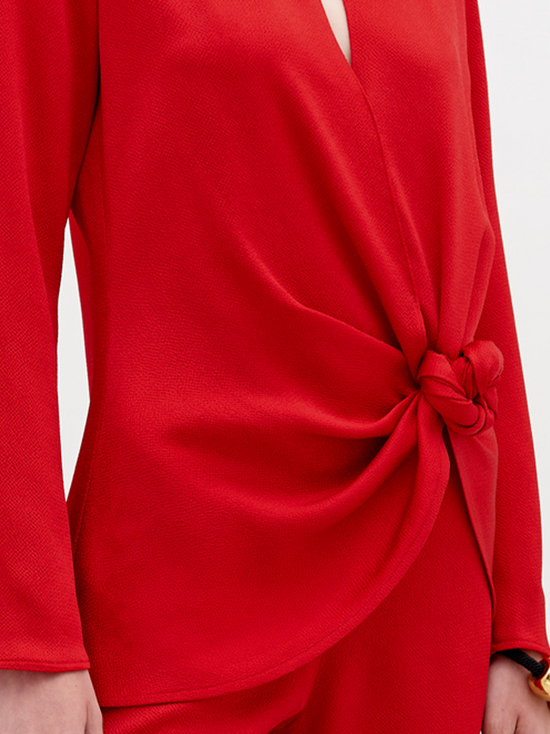 A woman stands in a red outfit with a knot detail on the top. She has her hair pulled back and stares directly into the camera against a plain white background. Available for pre-order now, the Tayra Blouse Rouge will have a ship date of November 15th, 2024.