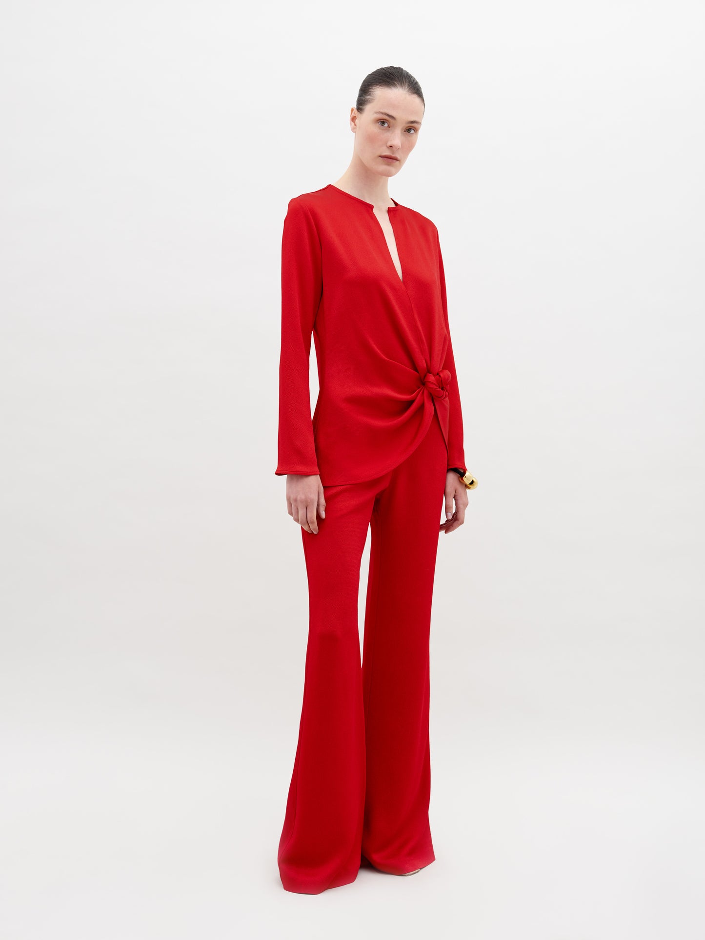 A person stands against a plain background wearing a red long-sleeved top with a knot detail at the waist and matching Palermo Pant Rouge, available for pre-order with a ship by date of November 15th, 2024.