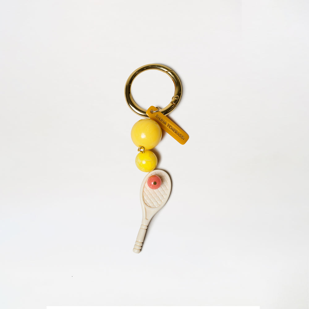 The Deuce Charm is a keychain featuring a gold ring, two yellow beads, an orange LOEWE FOUNDATION tag, and a miniature white tennis racket charm with an orange dot at its center.