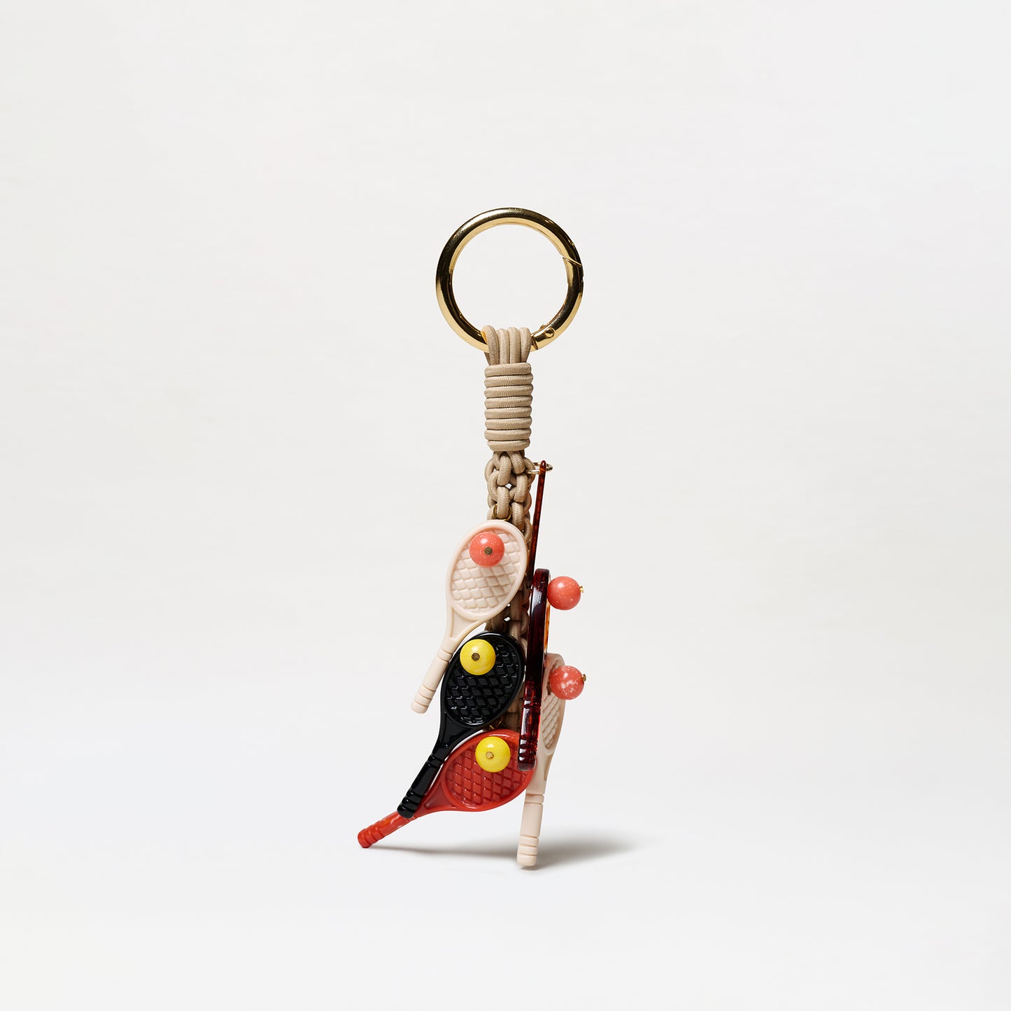 The Tournament Charm is a keychain with a gold ring, beige woven cord, and charms shaped like mini tennis rackets and balls in red, black, and yellow.