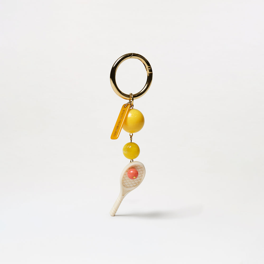 The Deuce Charm keychain features a gold ring, two yellow tennis ball beads, and a white tennis racket charm with a pink ball in the center, all displayed against a plain white background.